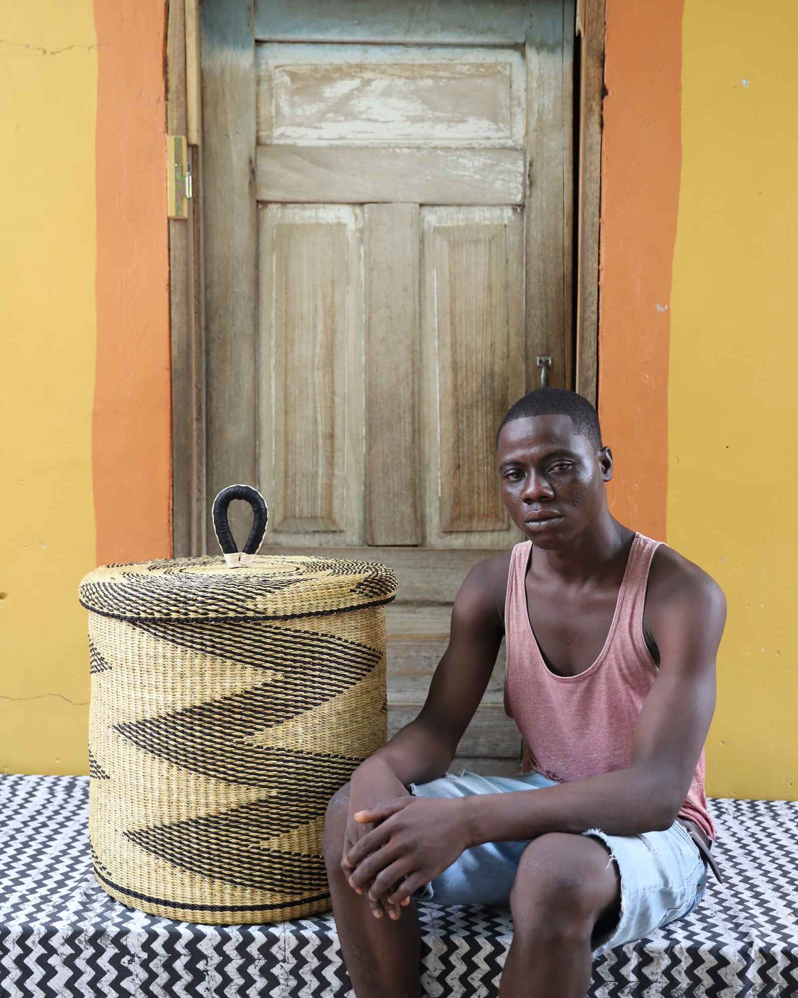 Handwoven Bolga laundry basket with lid in natural straw with a black zigzag pattern, styled beside a seated man.