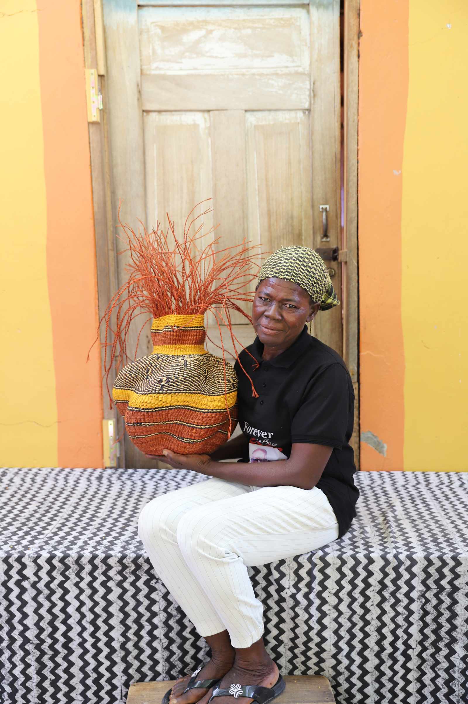 Handwoven Bolga Baobab basket in natural straw with orange band and grass sprigs, held by a seated woman.