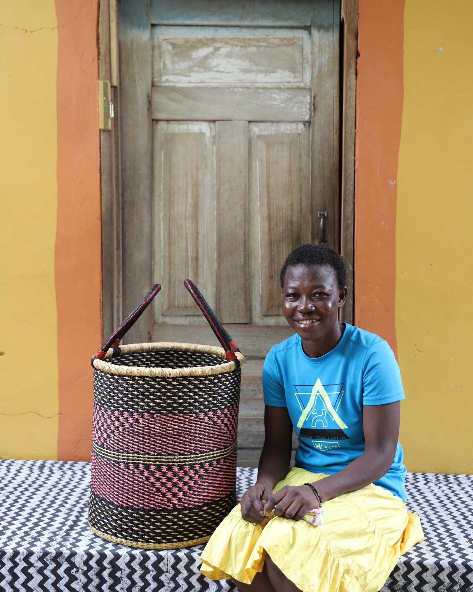 Handwoven open-top Bolga laundry basket in natural straw with black and red bands, styled beside a seated woman.
