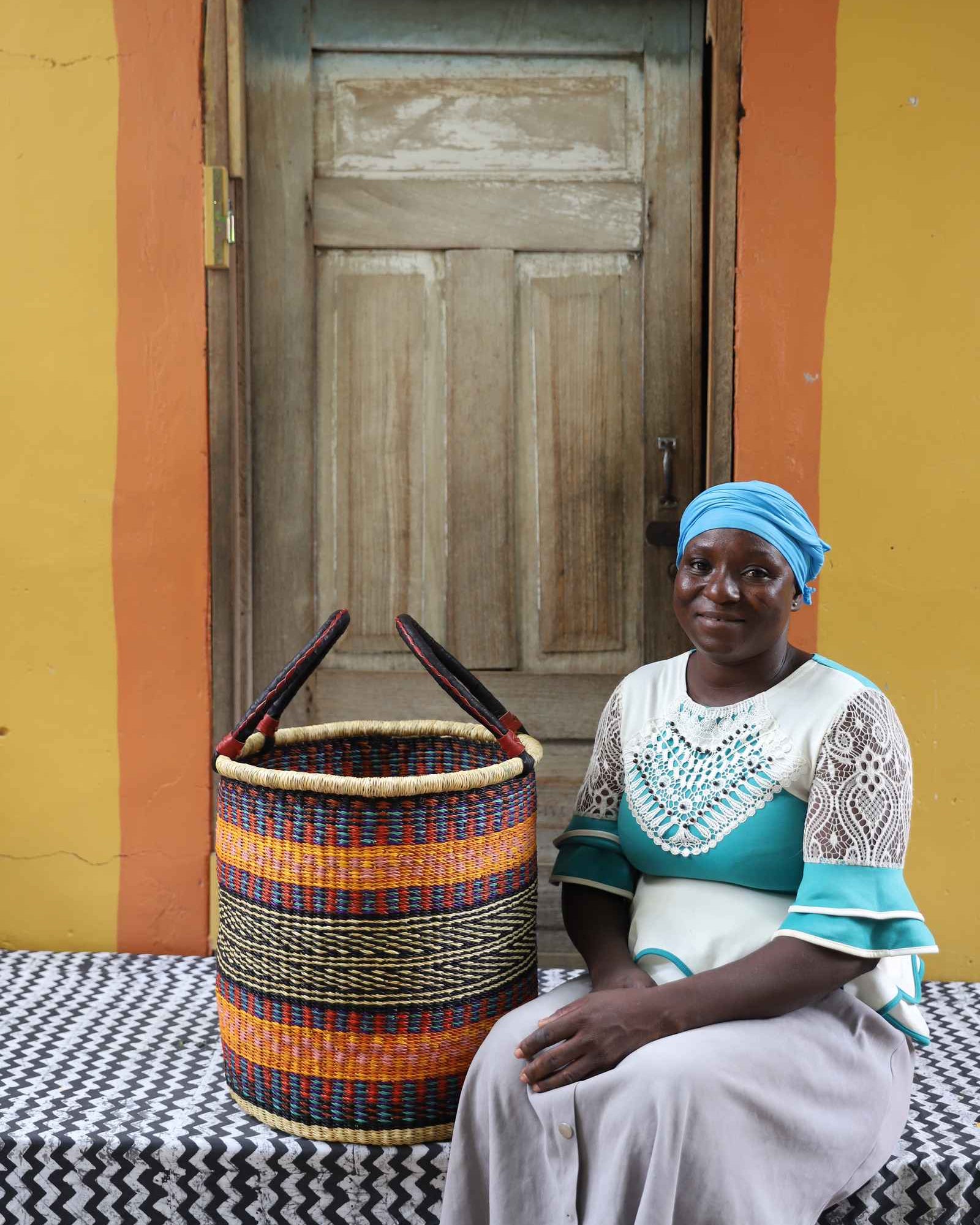Handwoven open-top Bolga laundry basket in natural straw with orange, yellow and black stripes, styled beside a seated woman.