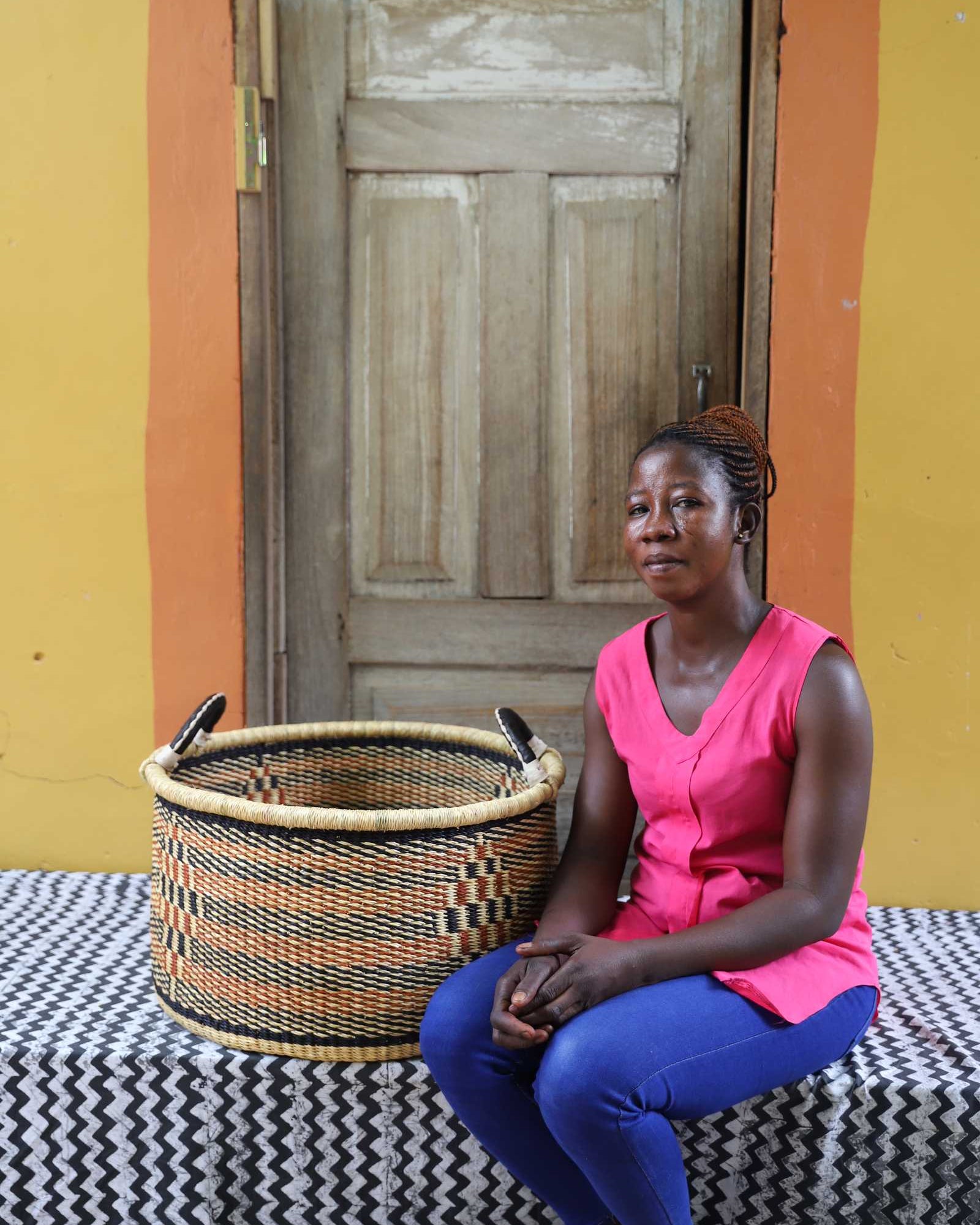 Handwoven Bolga storage basket in natural straw with orange and black stripes, styled beside a seated woman.