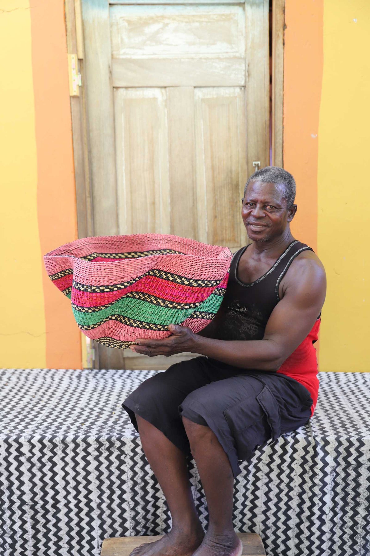 Handwoven Bolga wave basket in pink, green and black wavy stripes, held by a seated man in a black vest.