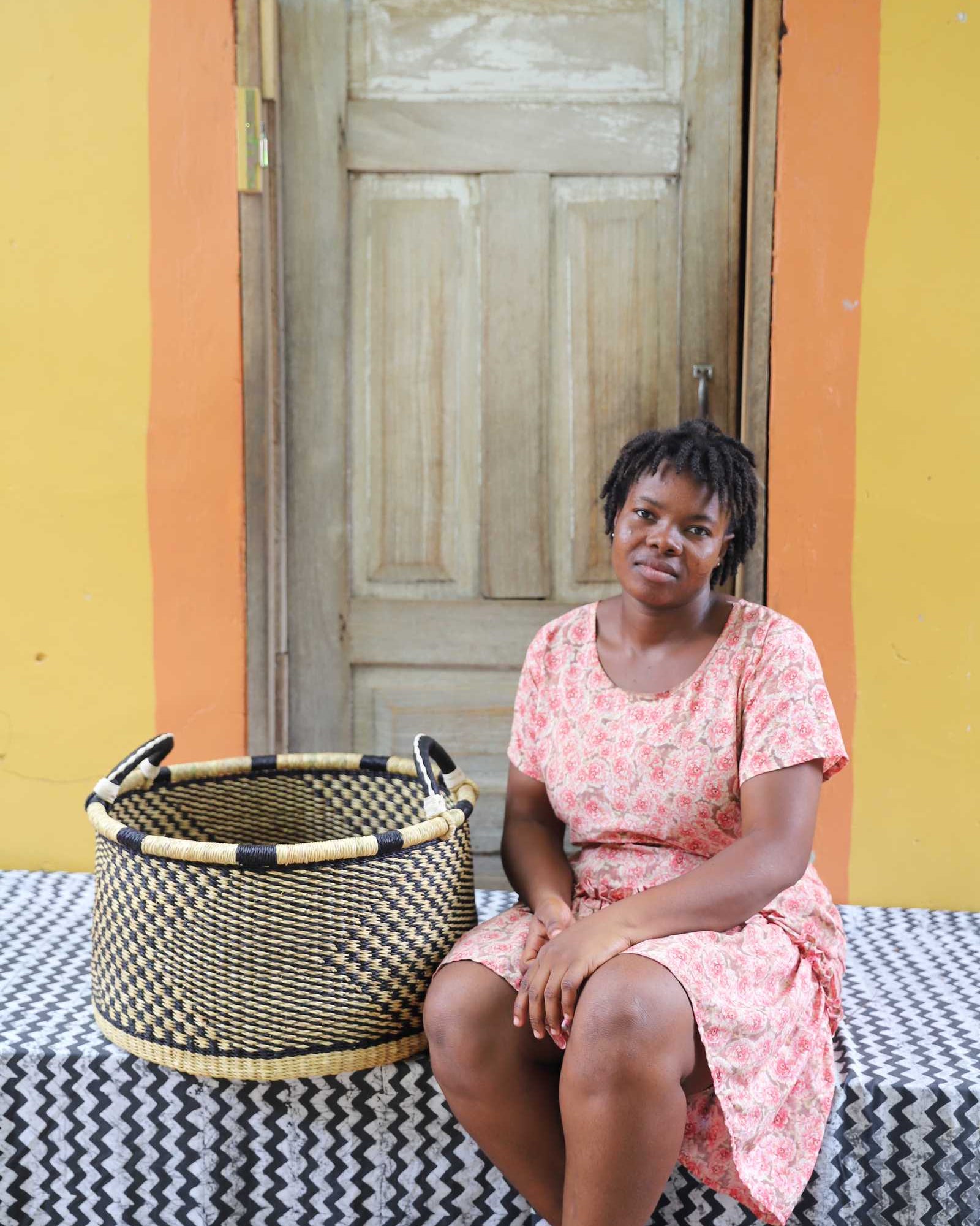 Handwoven Bolga storage basket in natural straw with a black geometric weave, styled beside a seated woman.