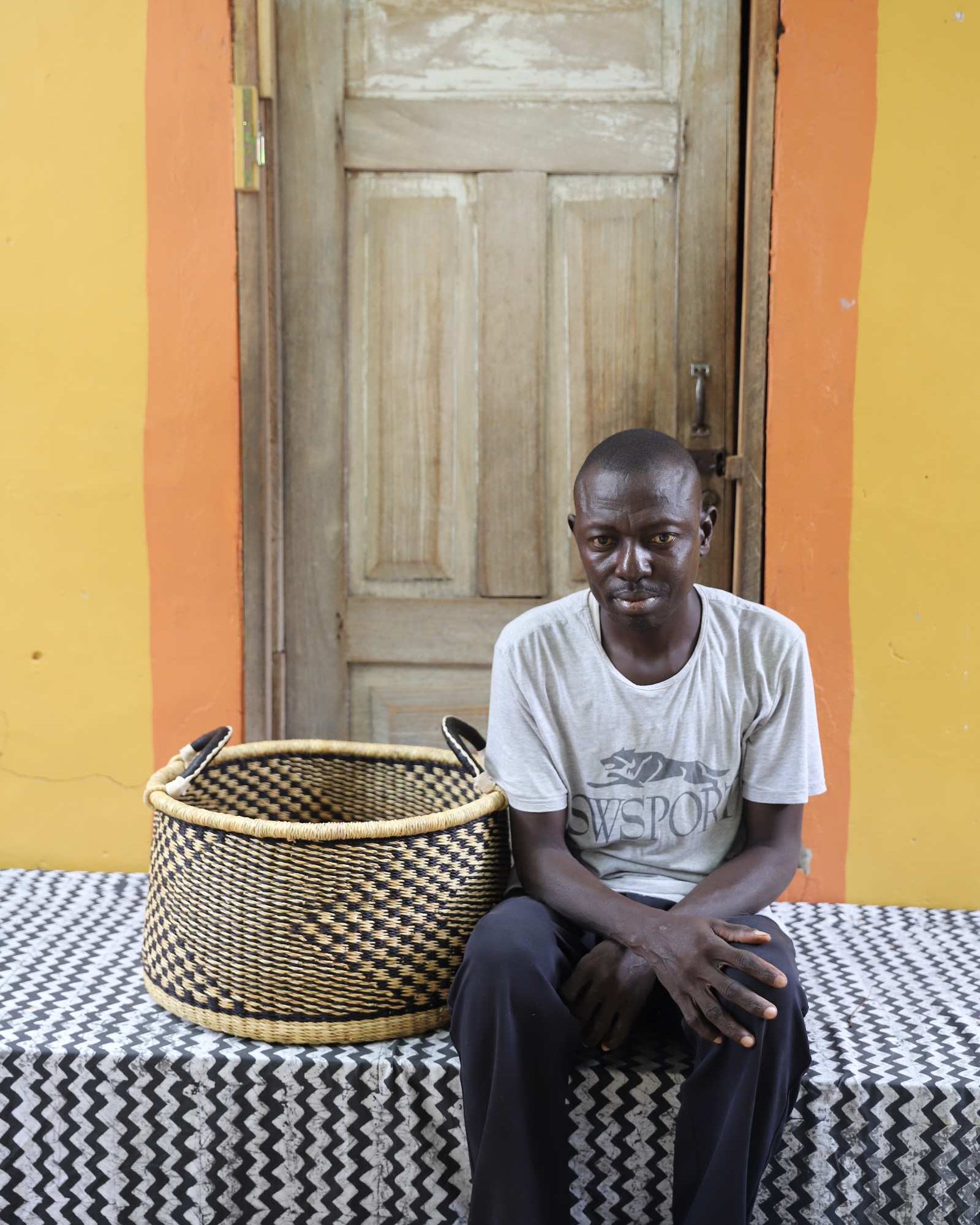 Handwoven Bolga storage basket in natural straw with a dark geometric weave, styled beside a seated man.