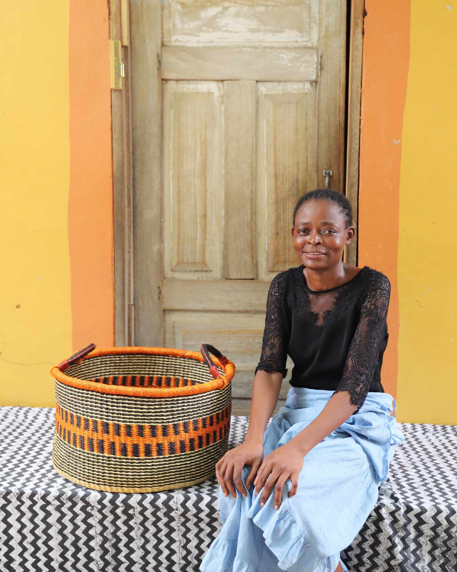 Handwoven Bolga storage basket in natural straw with an orange striped band, styled beside a seated woman.