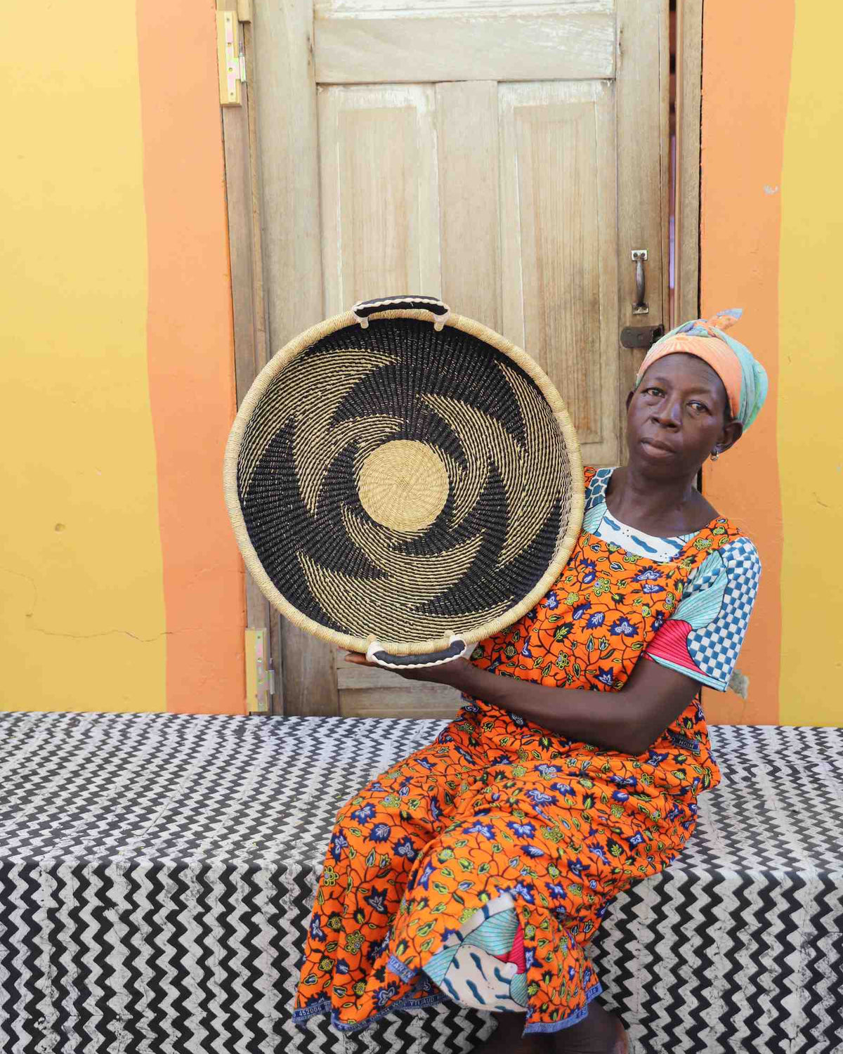 Handwoven Bolga wall tray disc with dark pinwheel swirl, held by a seated woman in an orange dress.