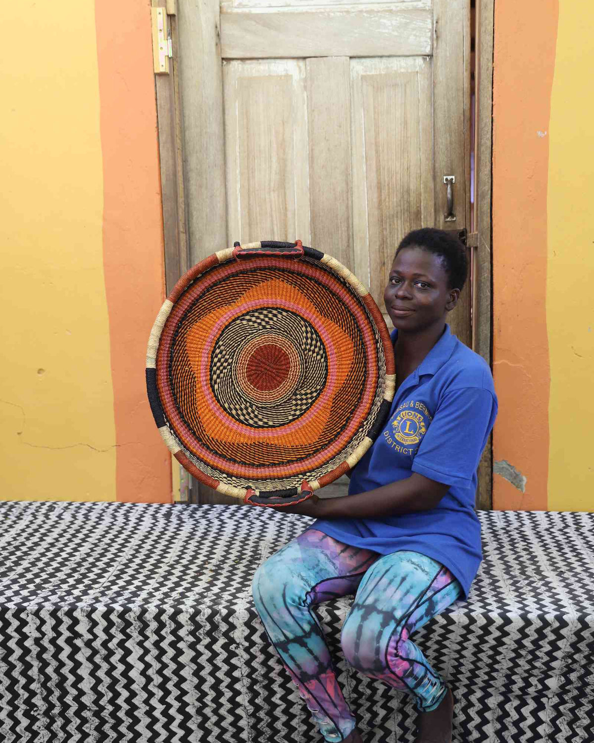Handwoven Bolga wall tray disc in rust-orange with black rings, held by a seated woman in blue.