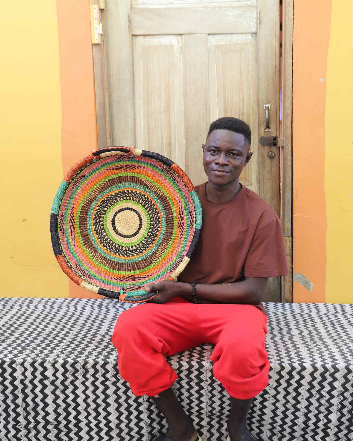 Handwoven Bolga wall tray disc with concentric multicolour rings, held by a seated man in red.