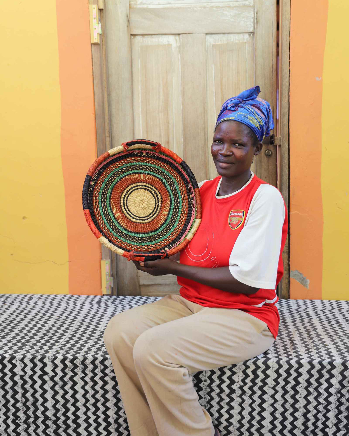 Handwoven Bolga wall tray disc in orange, black, red and green bands, held by a seated woman.