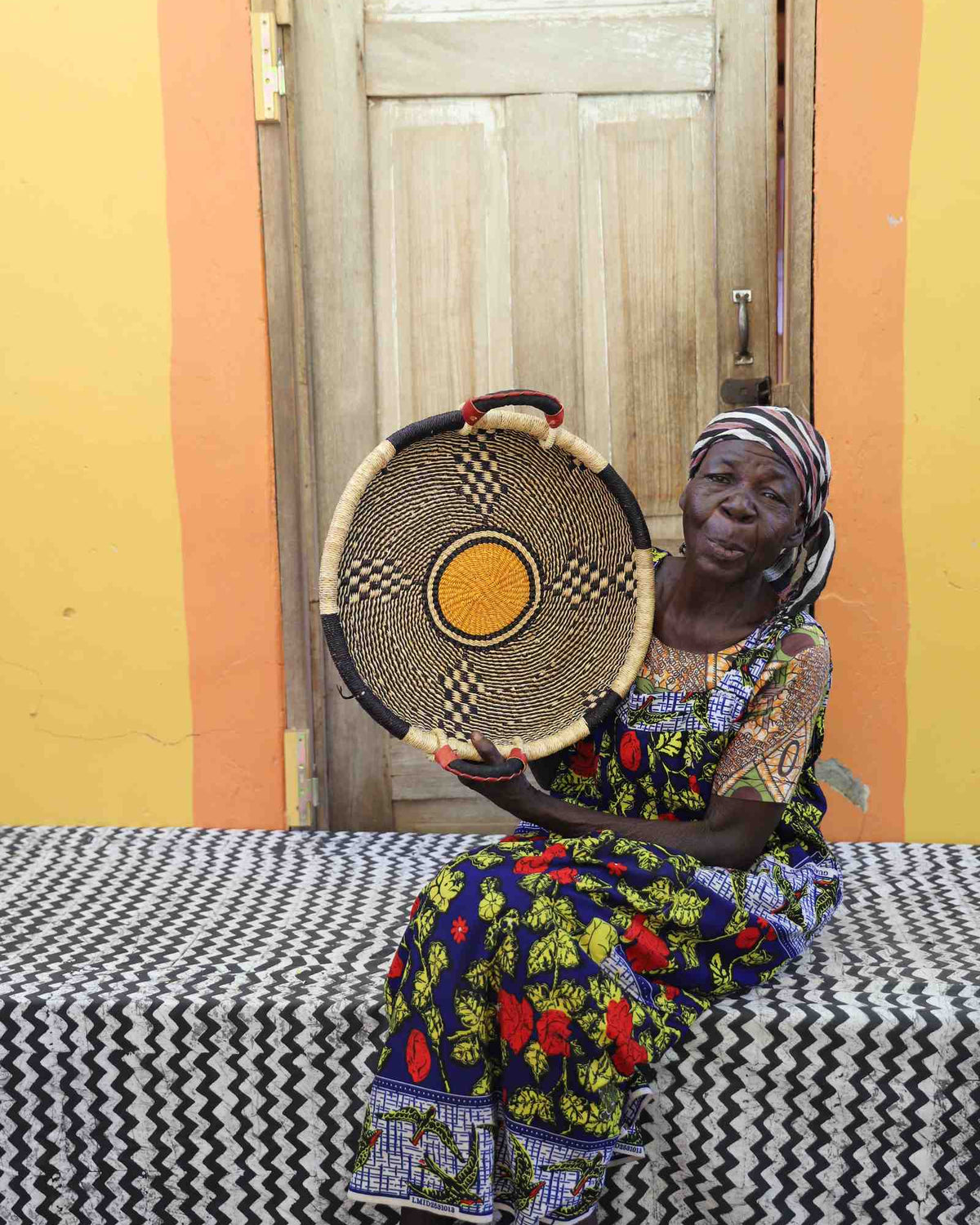 Handwoven Bolga wall tray disc in natural straw with a single yellow centre dot, held by a seated woman.