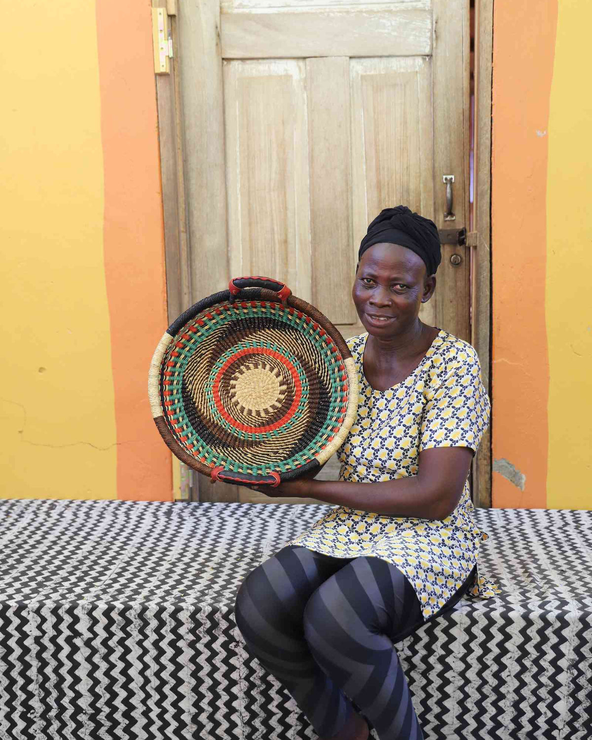 Handwoven Bolga wall tray disc with concentric pink, red and teal bands and yellow centre, held by a seated woman.