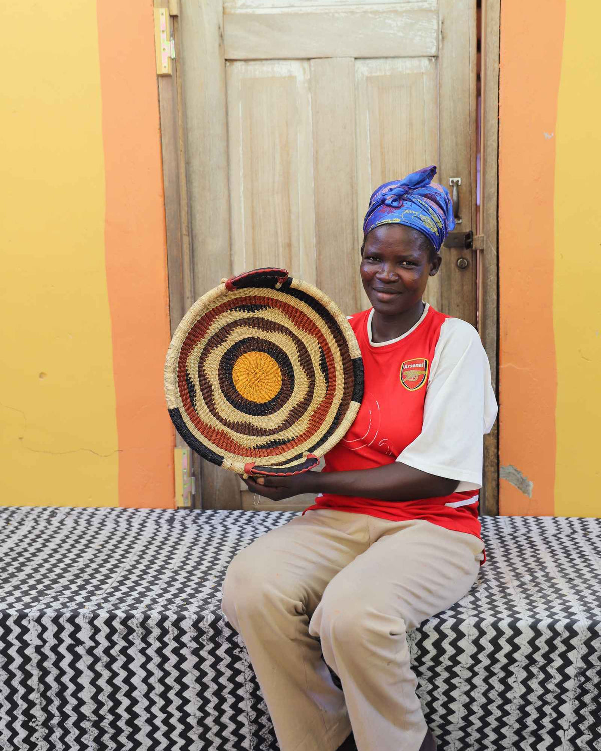 Handwoven Bolga wall tray disc with red, black and orange concentric bands, held by a seated woman.