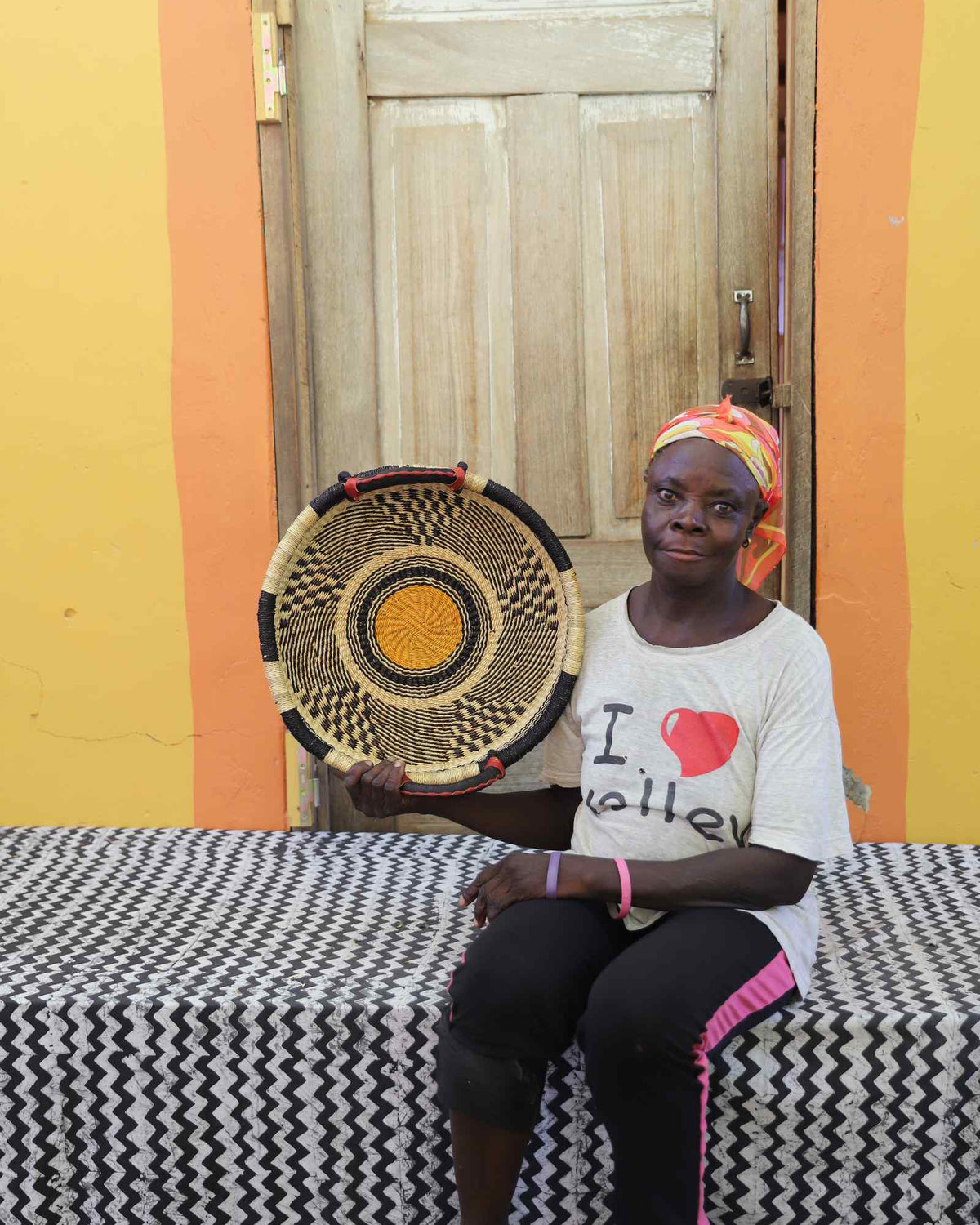 Handwoven Bolga wall tray disc in natural straw with a yellow-orange bullseye centre, held by a seated woman.