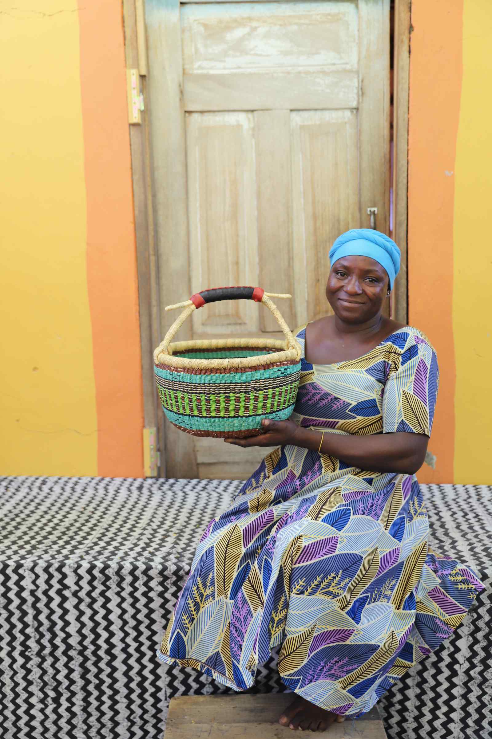 Handwoven round Bolga shopping basket in green and teal, held by a seated woman in a patterned dress.
