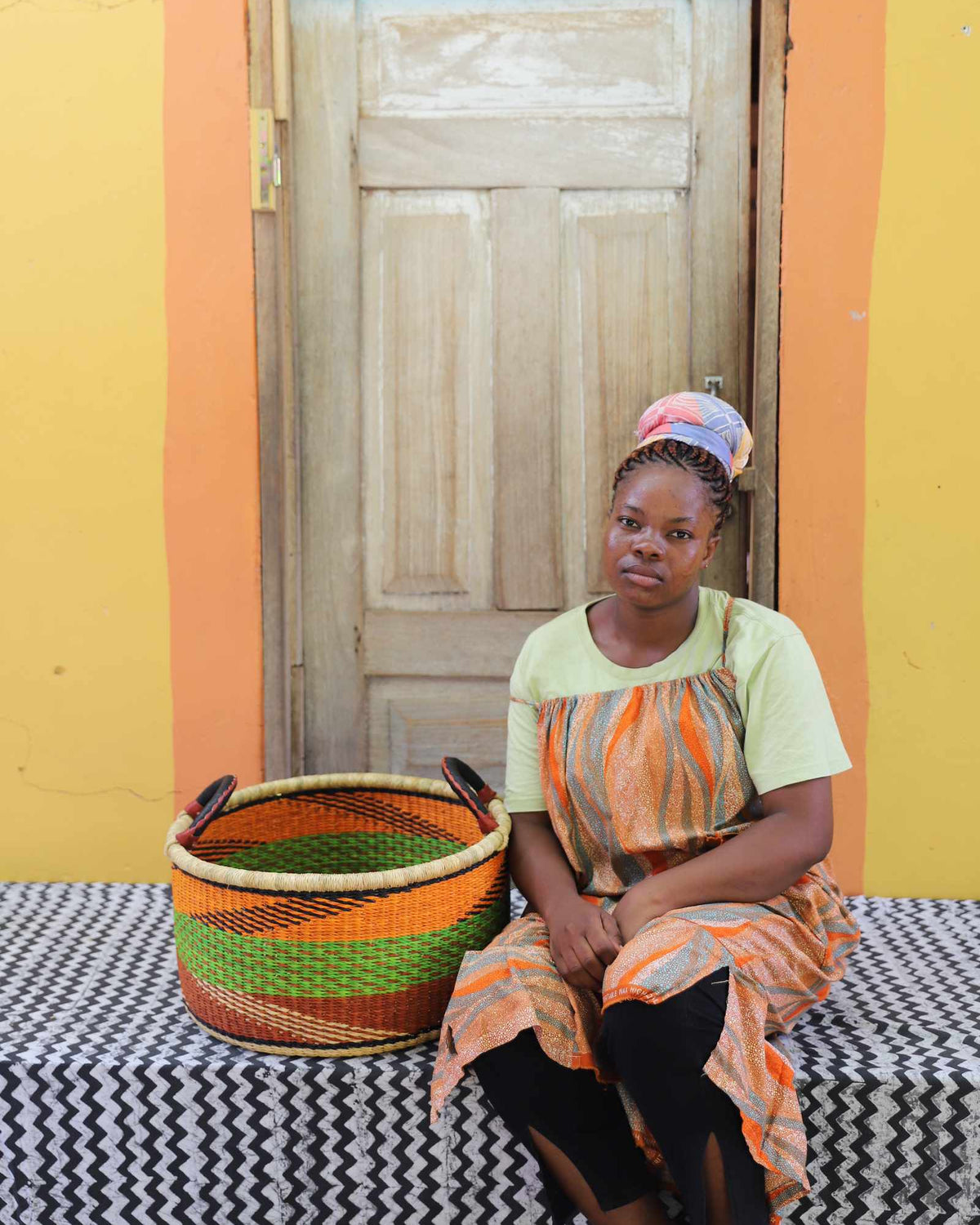 Small handwoven Bolga storage tub in natural with orange and green stripes, beside a seated woman in orange.