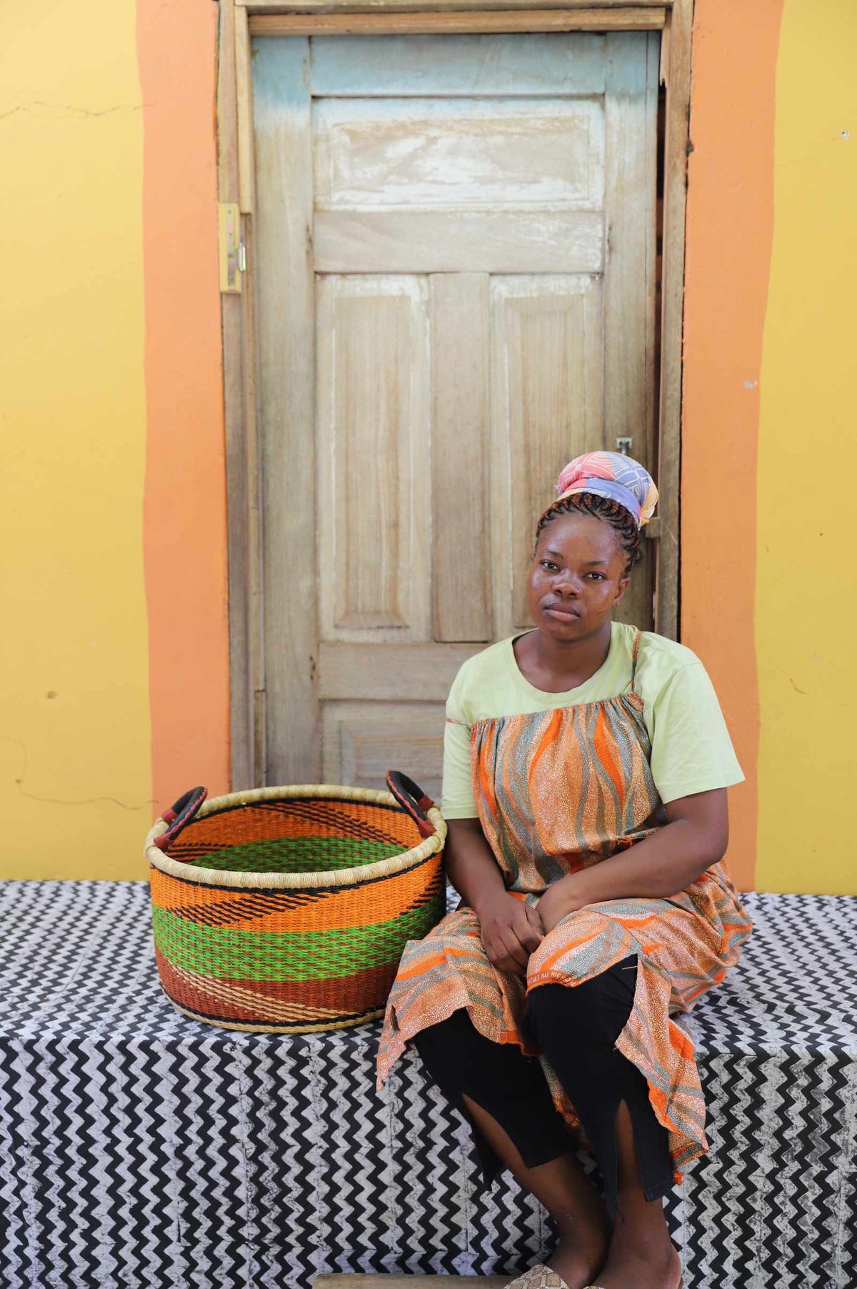 Small handwoven Bolga storage tub in natural with orange and green stripes, beside a seated woman in orange.