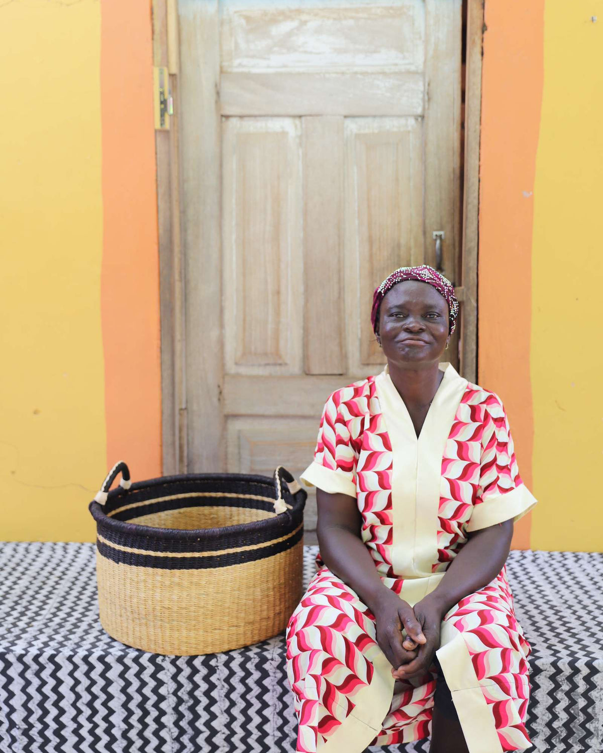 Small handwoven Bolga storage tub in plain natural straw, beside a seated woman in red and white.