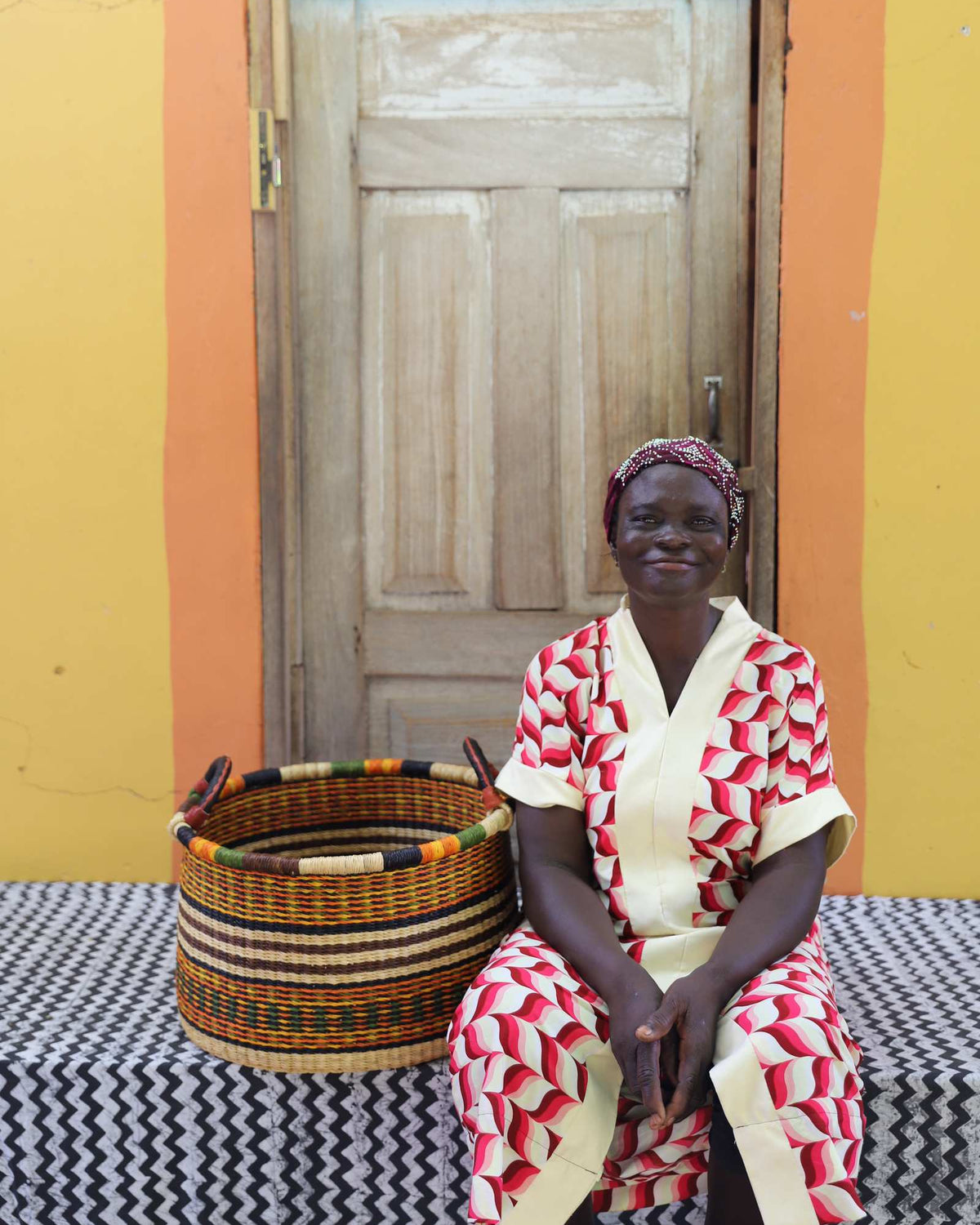Small handwoven Bolga storage tub in natural with fine red stripes, beside a seated woman in red and white.