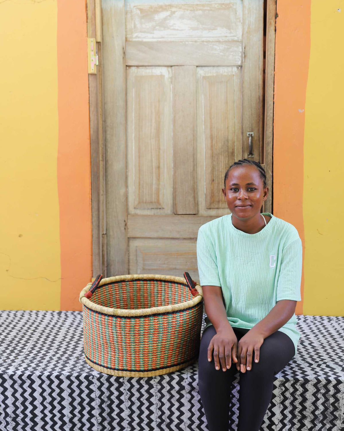 Small handwoven Bolga storage tub in natural with orange and green bands, beside a seated woman in green.
