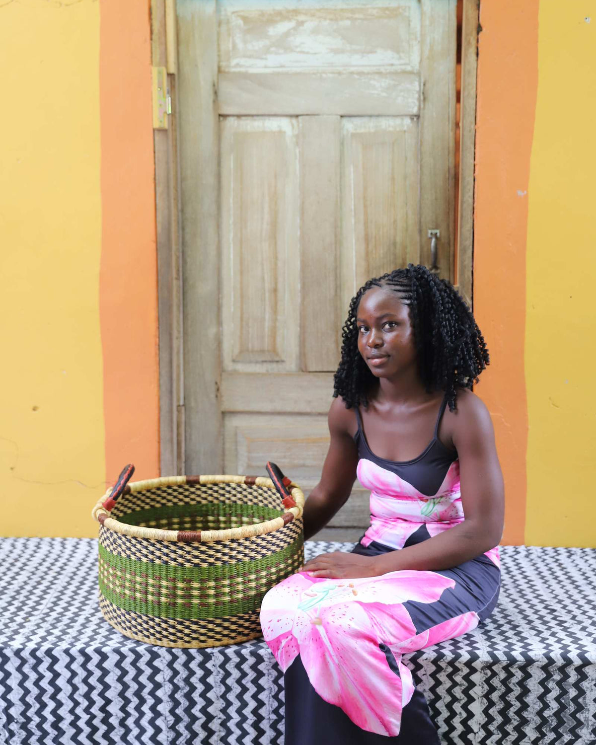Small handwoven Bolga storage tub in dark green, beside a seated woman in pink and black.