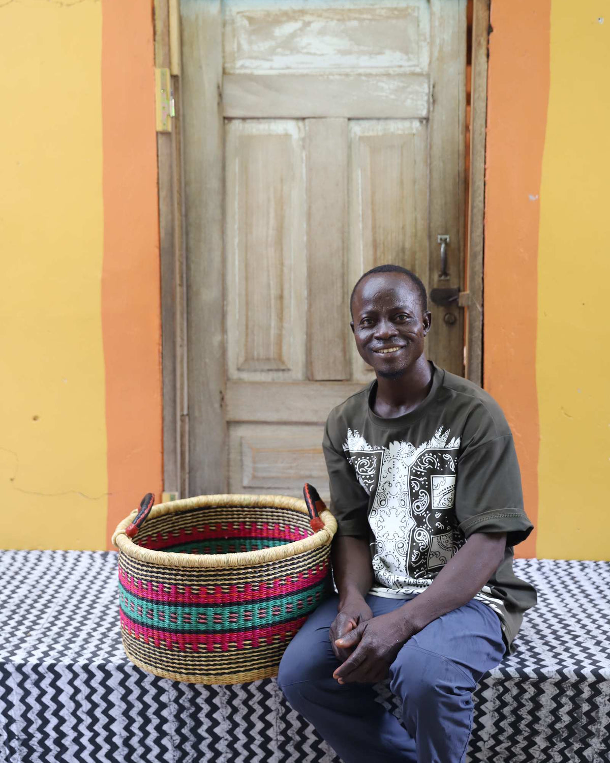 Small handwoven Bolga storage tub in natural with red and green bands, beside a seated man in patterned shirt.