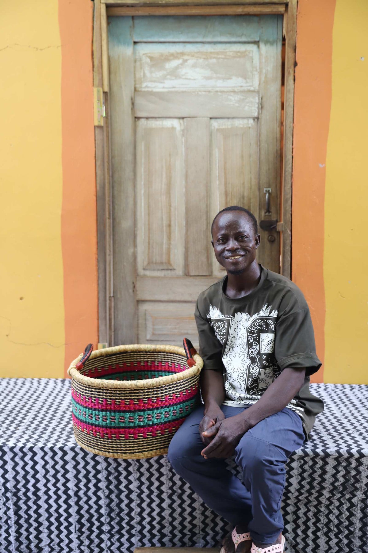 Small handwoven Bolga storage tub in natural with red and green bands, beside a seated man in patterned shirt.