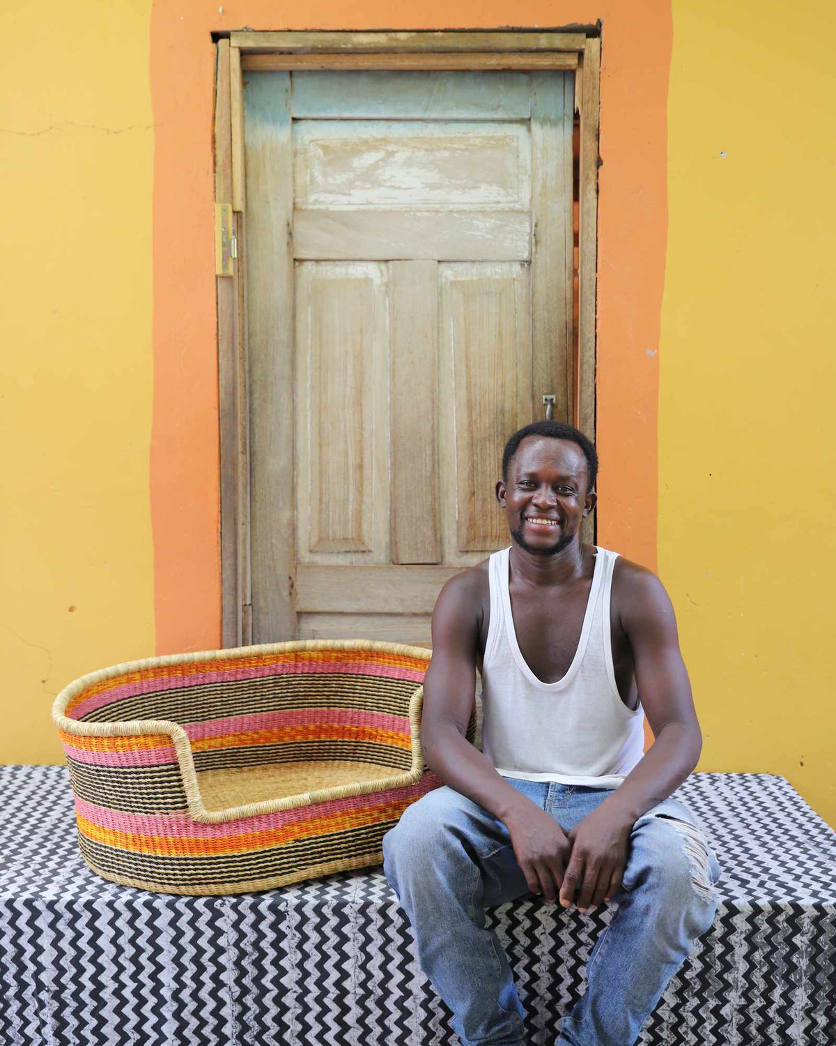 Large handwoven Bolga pet bed in natural with orange and red stripes, beside a seated man in a white vest.