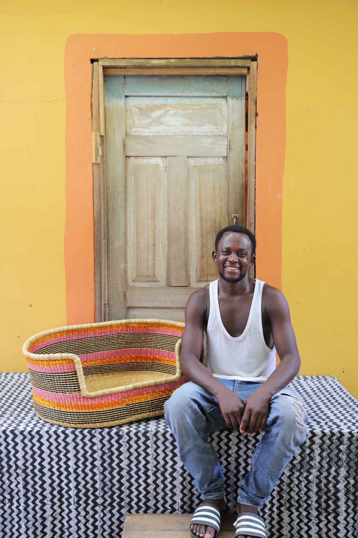 Large handwoven Bolga pet bed in natural with orange and red stripes, beside a seated man in a white vest.