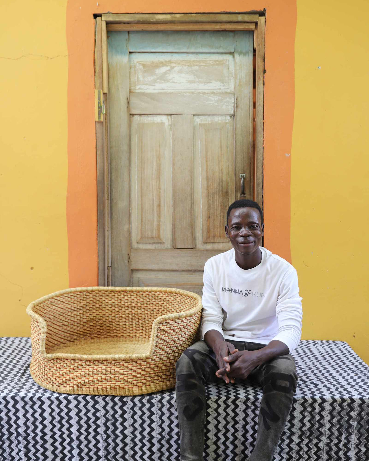 Large handwoven Bolga pet bed in plain natural straw, beside a seated man in white.