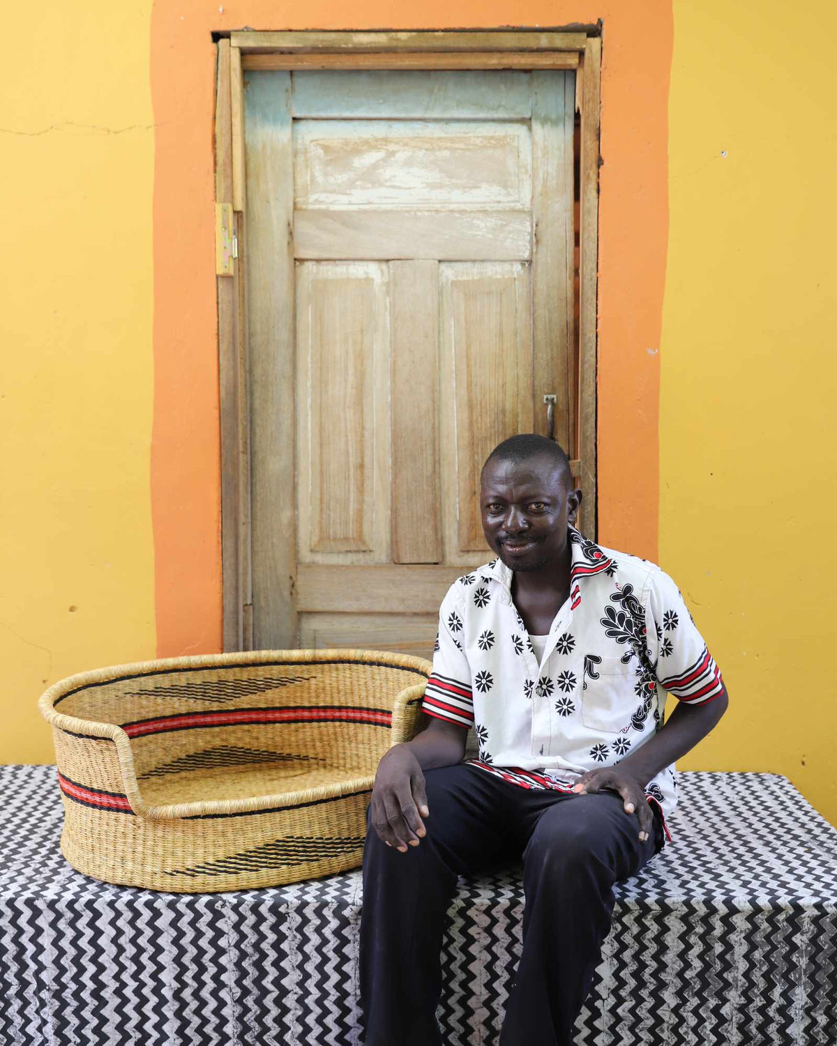 Large handwoven Bolga pet bed in natural with fine red stripes, beside a seated man in a printed shirt.