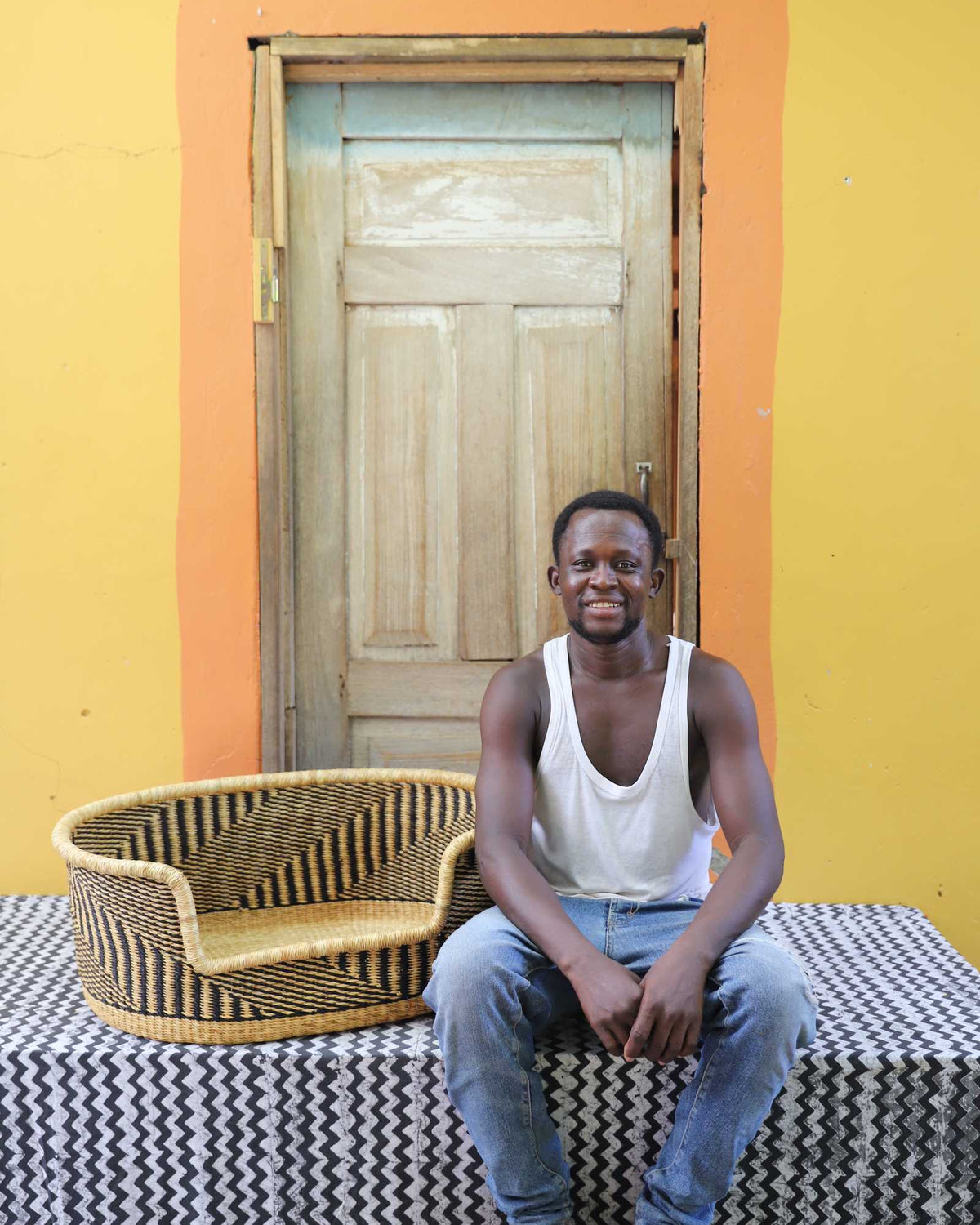 Medium handwoven Bolga pet bed in natural with black herringbone weave, beside a seated man in white.