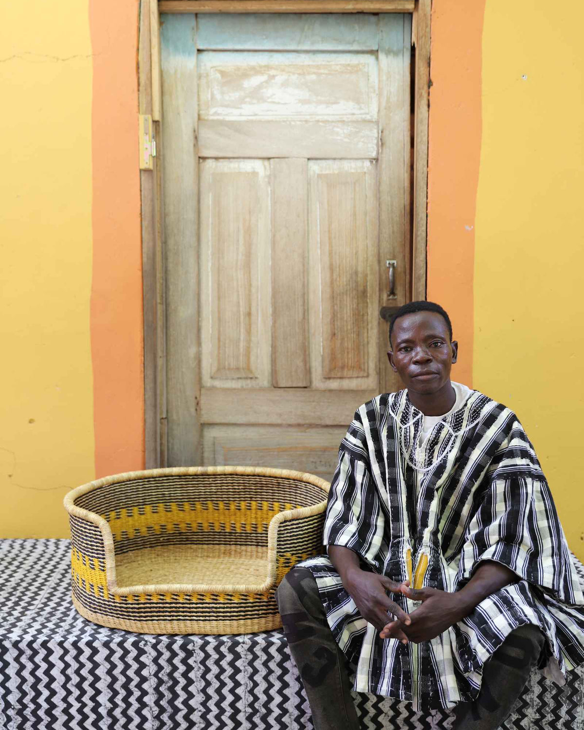 Medium handwoven Bolga pet bed in natural with yellow stripes, beside a seated man in stripes.