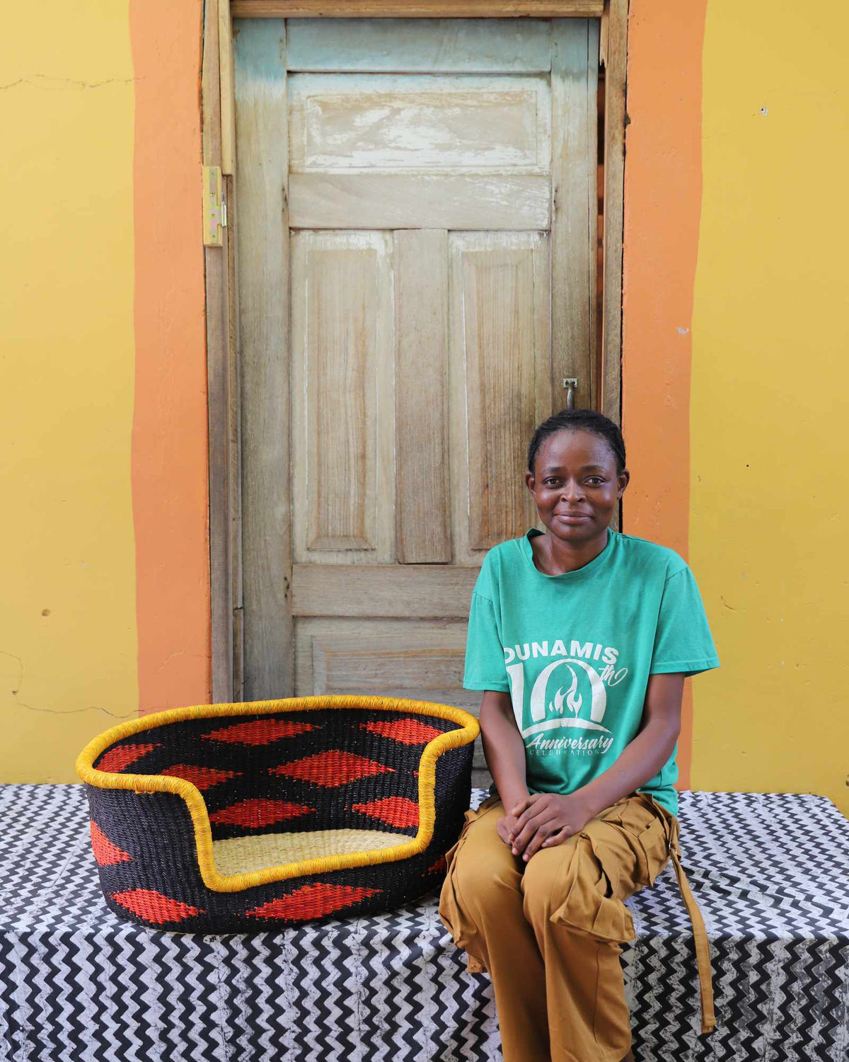 Small handwoven Bolga pet bed in black with orange diamonds, beside a seated woman in green.