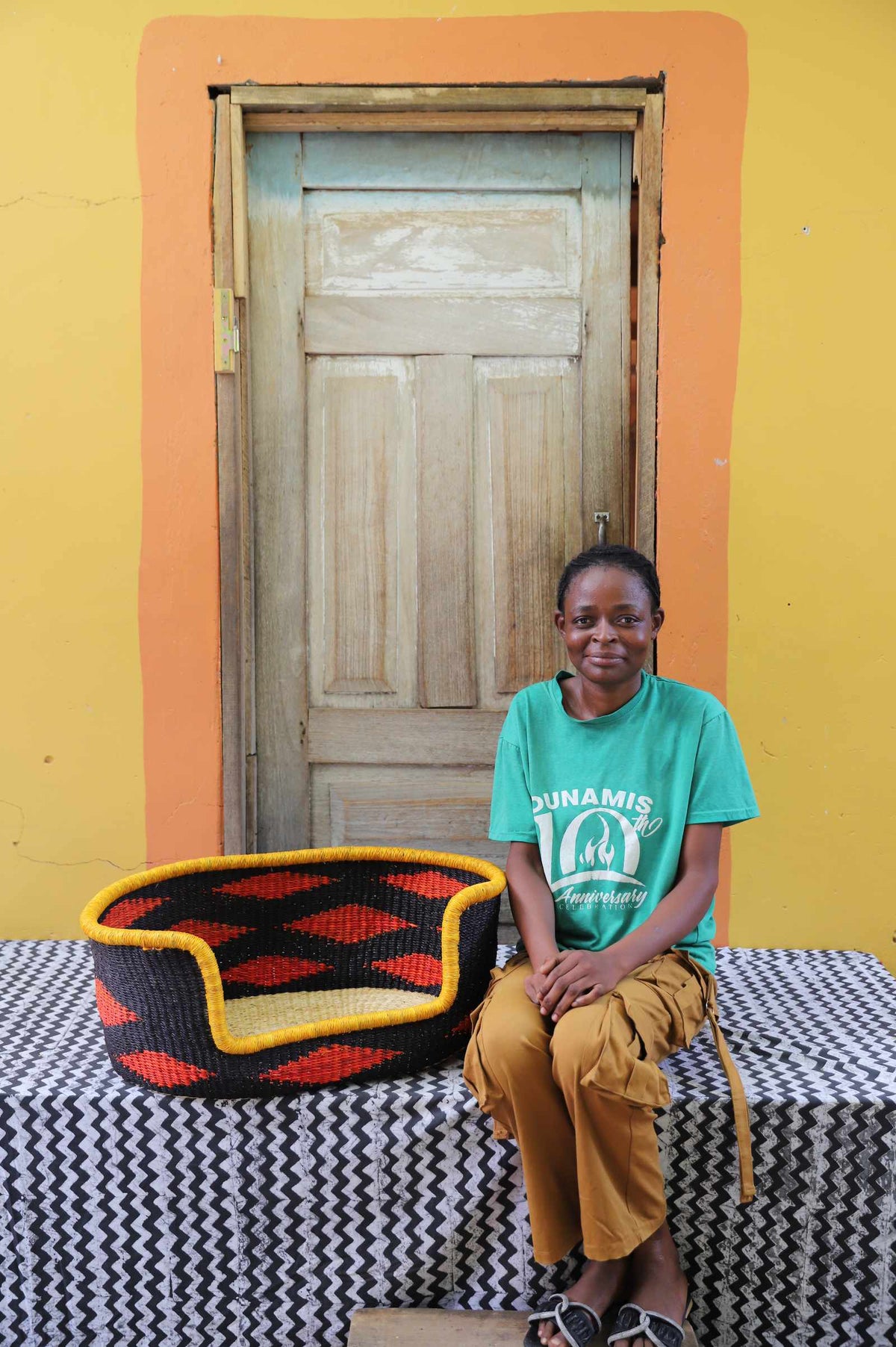 Small handwoven Bolga pet bed in black with orange diamonds, beside a seated woman in green.