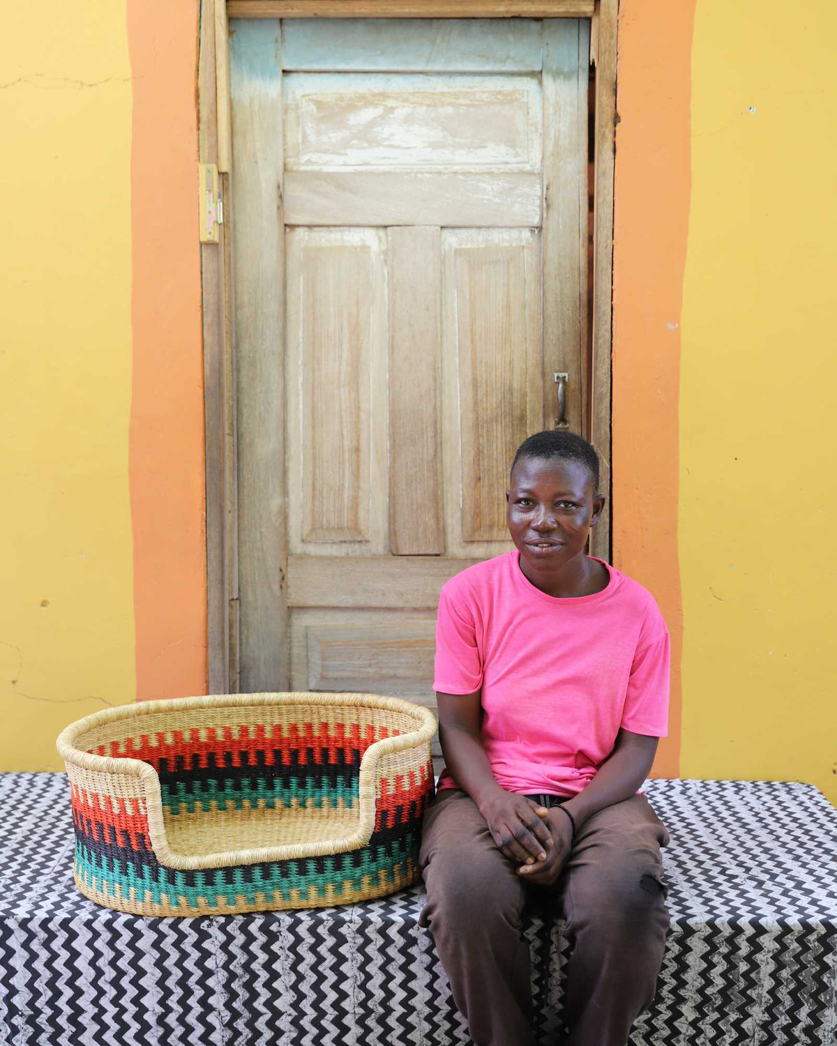 Small handwoven Bolga pet bed in natural with red, green and yellow stripes, beside a seated man in pink.