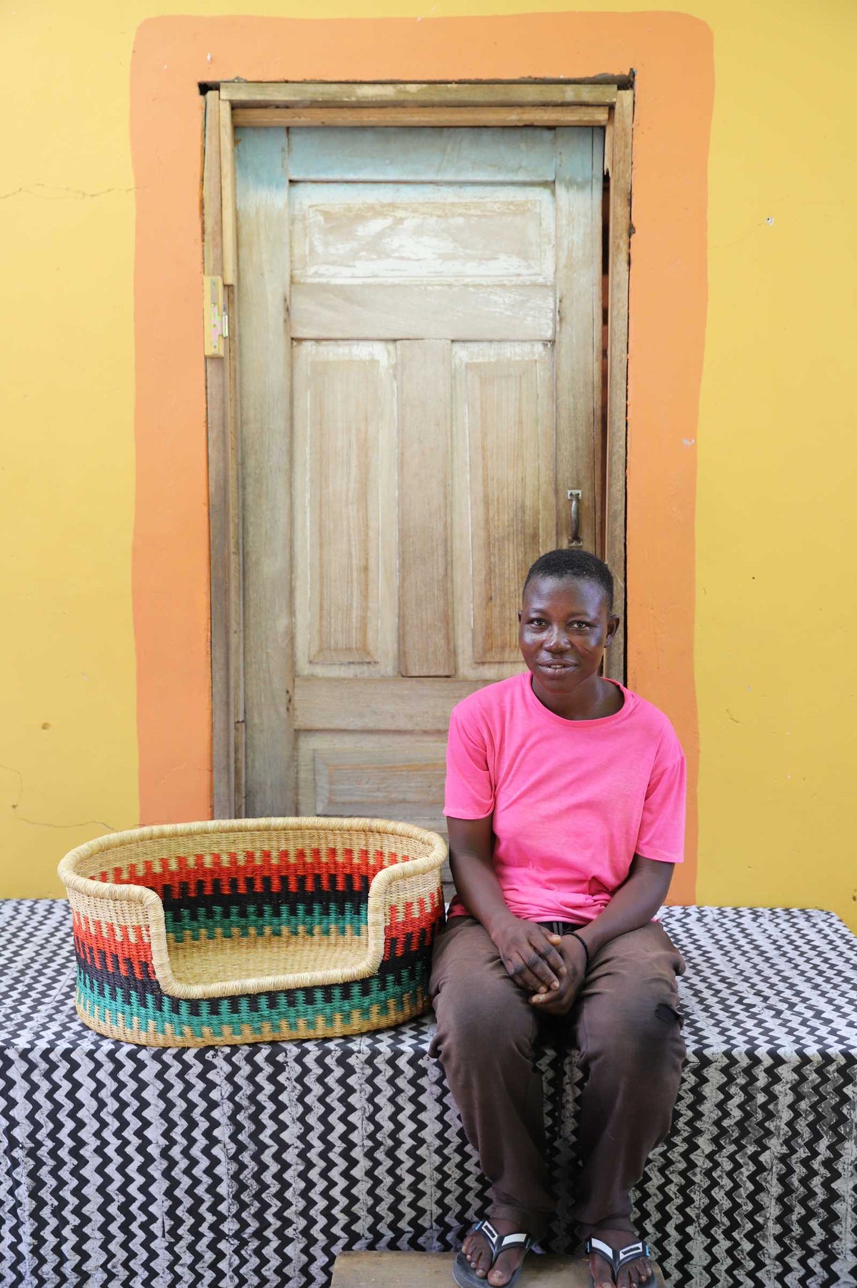 Small handwoven Bolga pet bed in natural with red, green and yellow stripes, beside a seated man in pink.