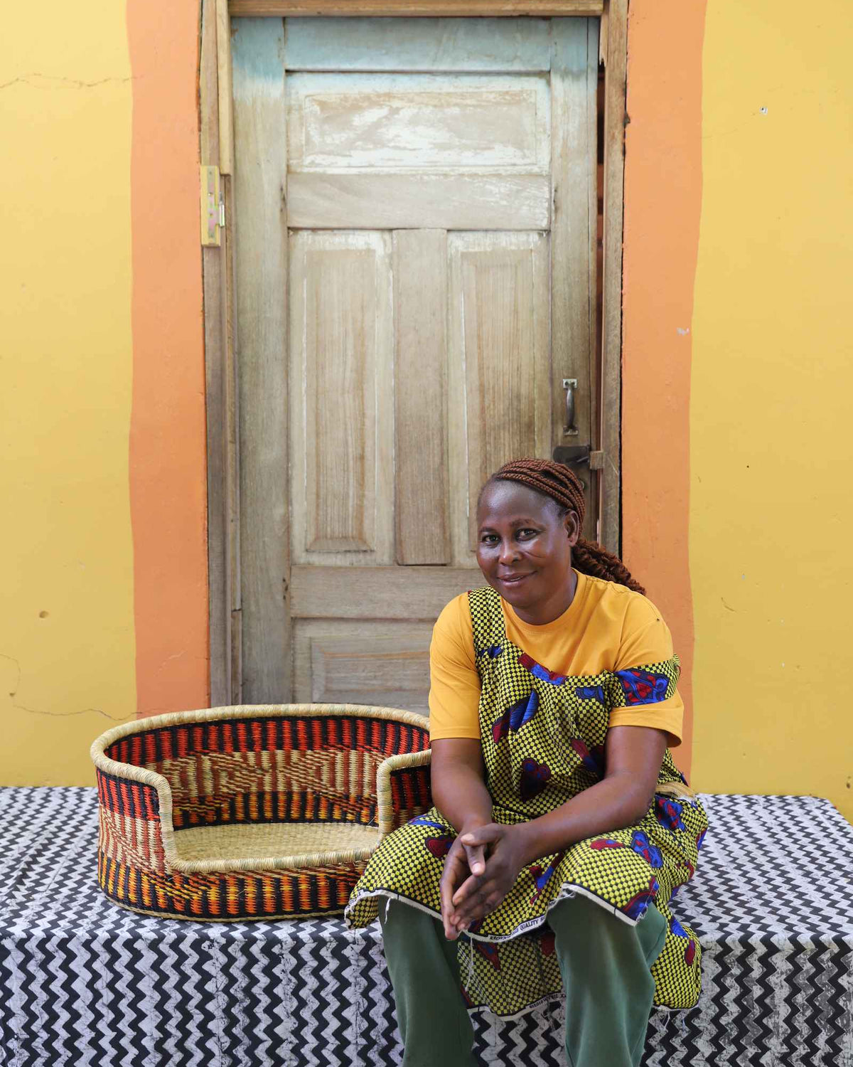 Small handwoven Bolga pet bed in orange, red and black stripes, beside a seated woman in yellow.