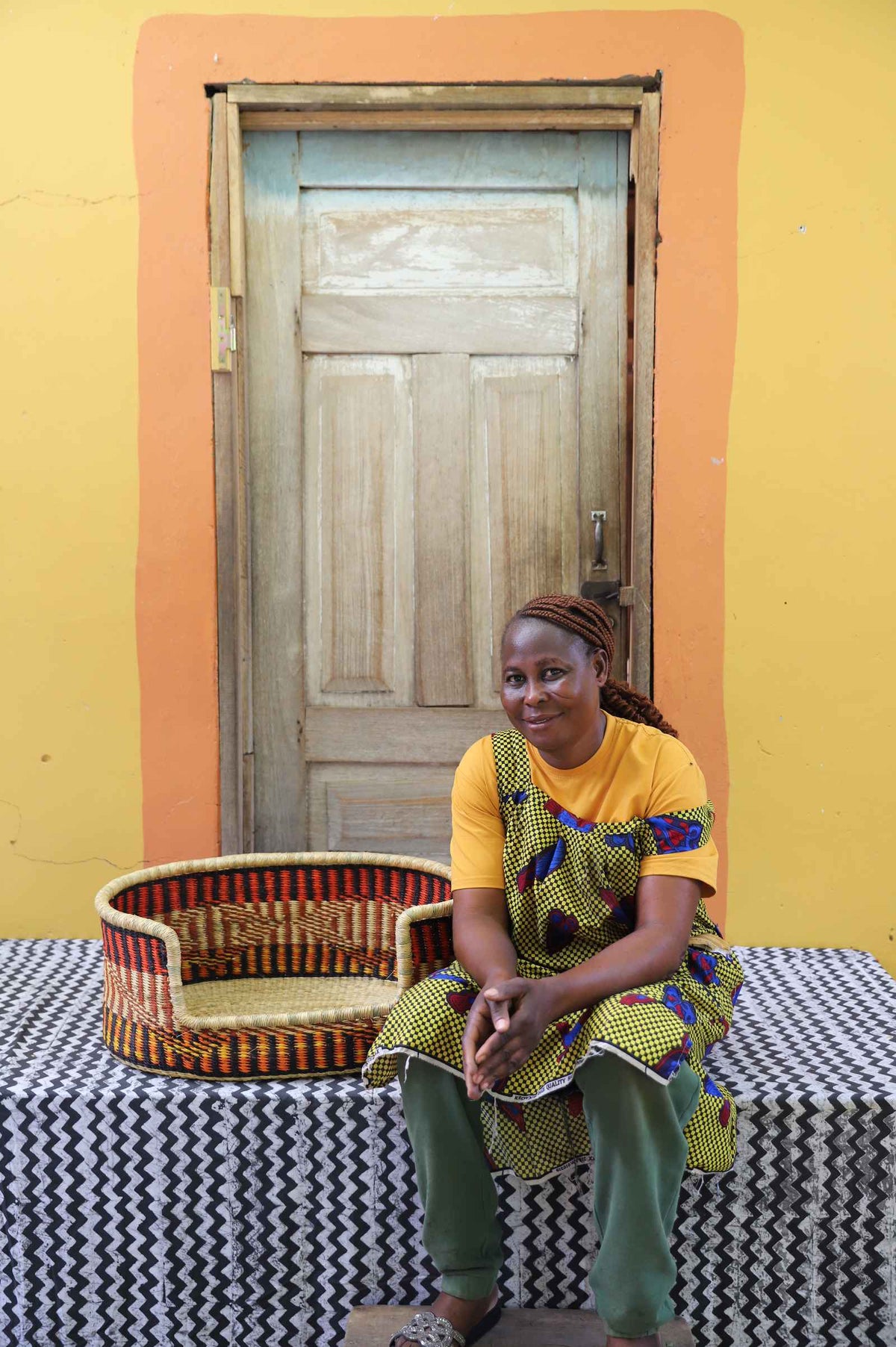Small handwoven Bolga pet bed in orange, red and black stripes, beside a seated woman in yellow.