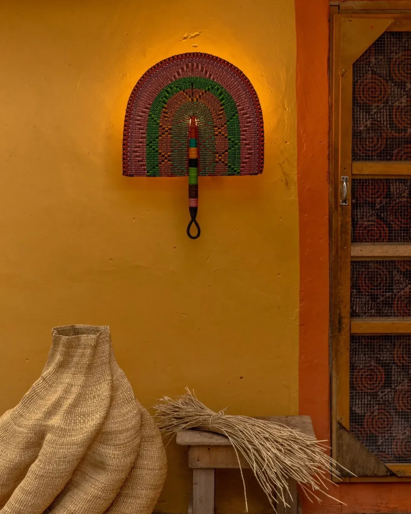 Luna wall lampshade by The Baba Tree, handwoven elephant grass (straw) shade shown beside a colourful wall and a beautiful sculptural basket with bundle of straw