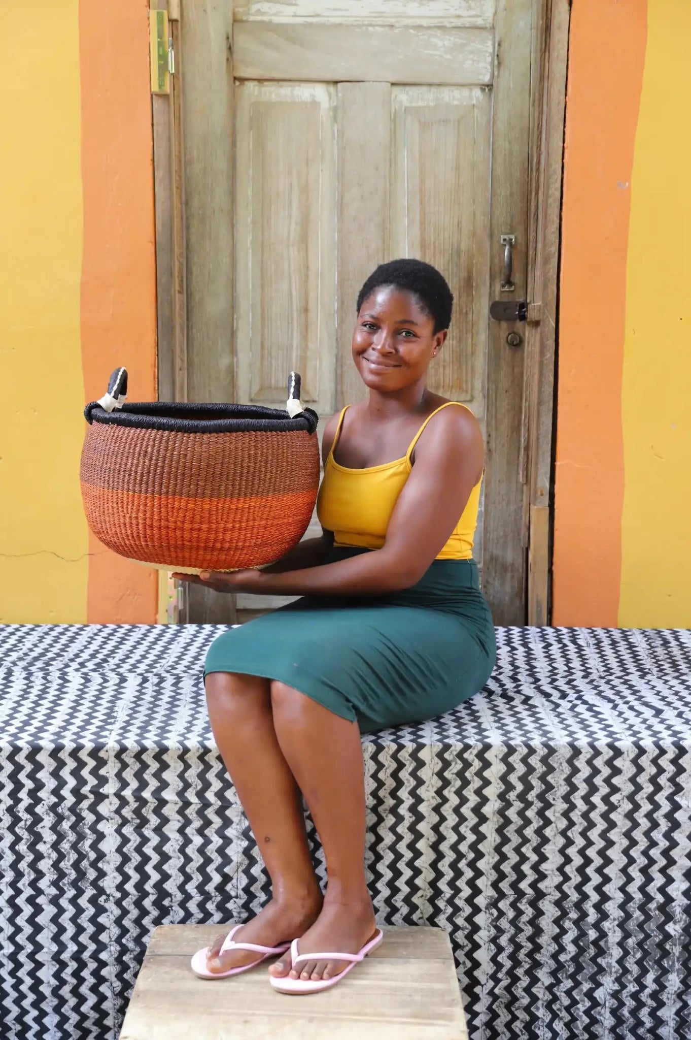 Artisan holding a colorful handwoven storage basket sitting on a black and white cloth against a wooden door.