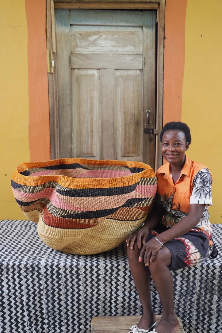 Woman sitting next to a large woven basket with geometric pattern on a black and white checkered tablecloth in front of a wooden door.