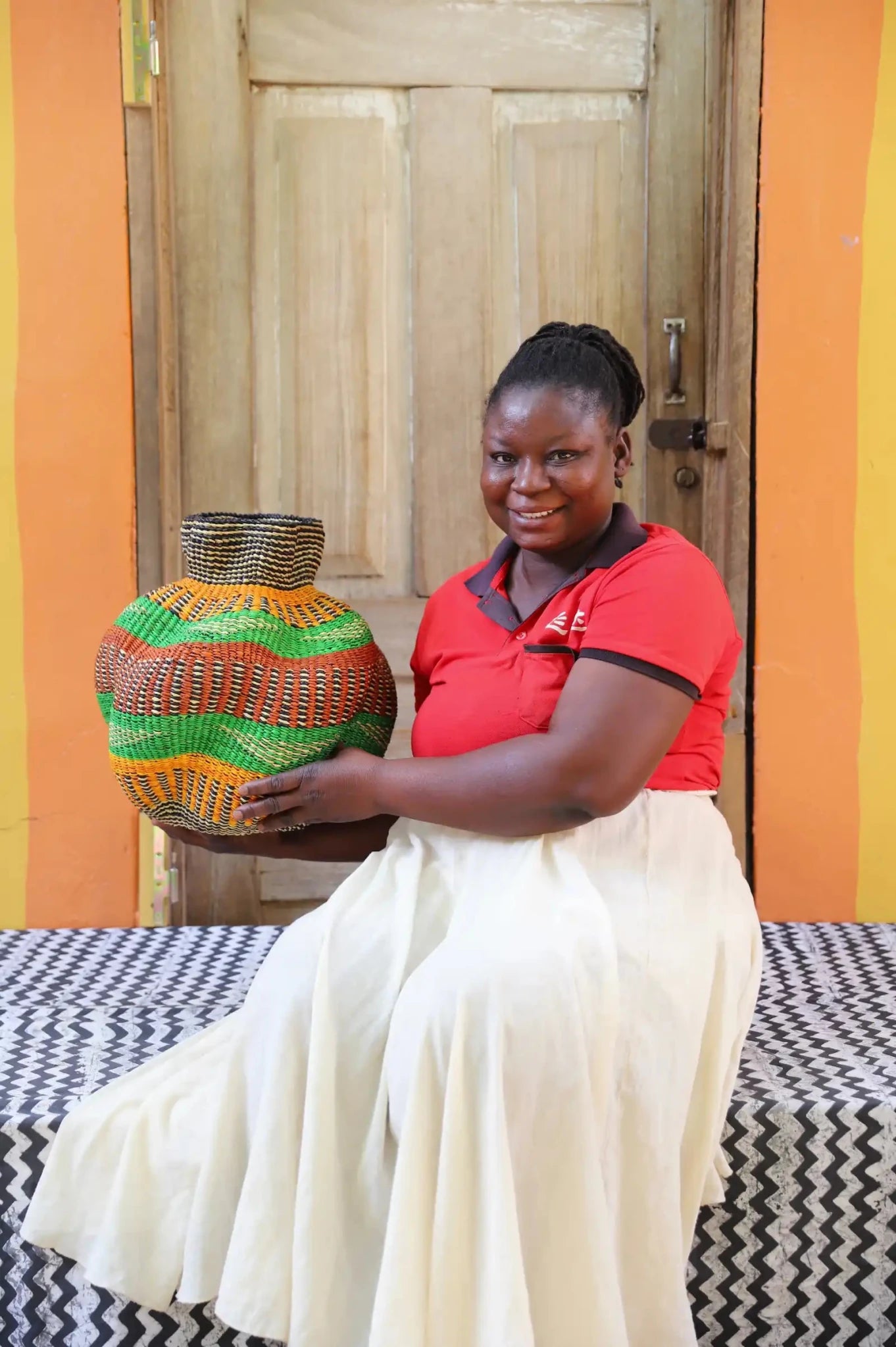 An artisan seated next to a handcrafted decorative basket  on a black and white cloth against a wooden door.