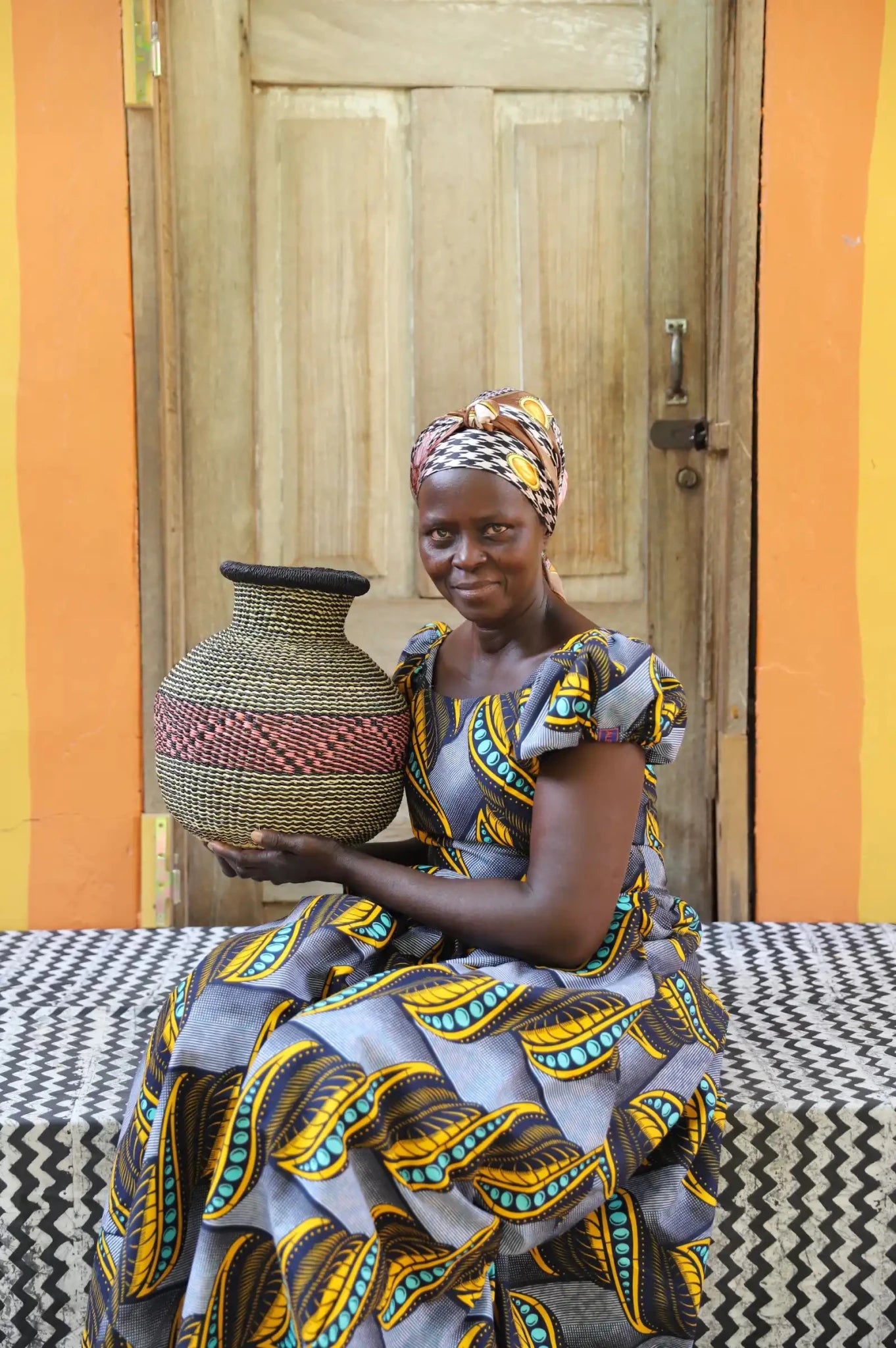 Artisan holding a colorful woven storage basket  on a black and white cloth against a wooden door.