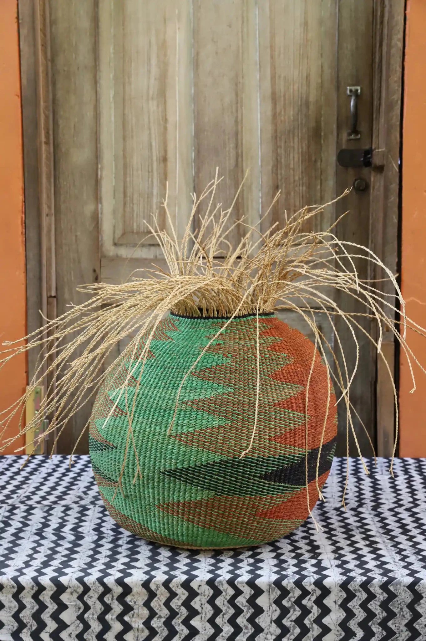 Decorative woven basket with stylish straw branching out on a black and white cloth in front of a wooden door.