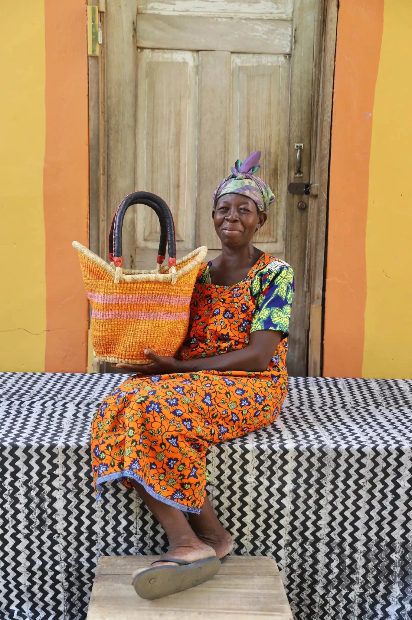 Woman in orange floral dress and headwrap holding a small Solara Carryall with natural and pink stripe