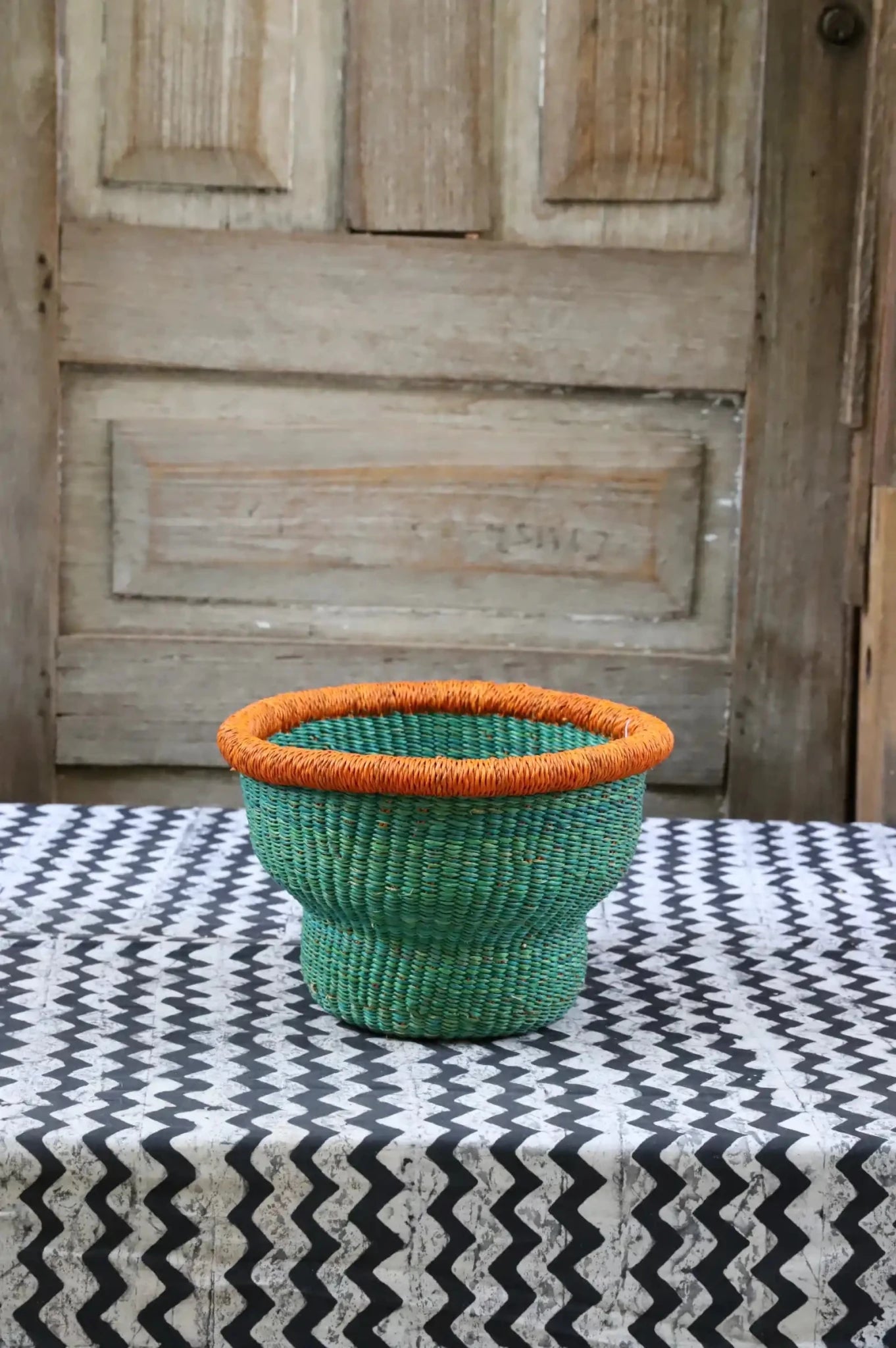 Green woven basket with orange rim on a black and white patterned tablecloth in front of a wooden door.