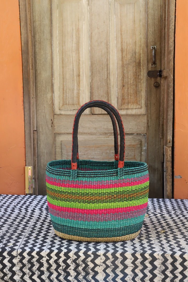 Round carry basket with teal, lime green and pink horizontal stripe bands, dark leather handles, against a wooden door.
