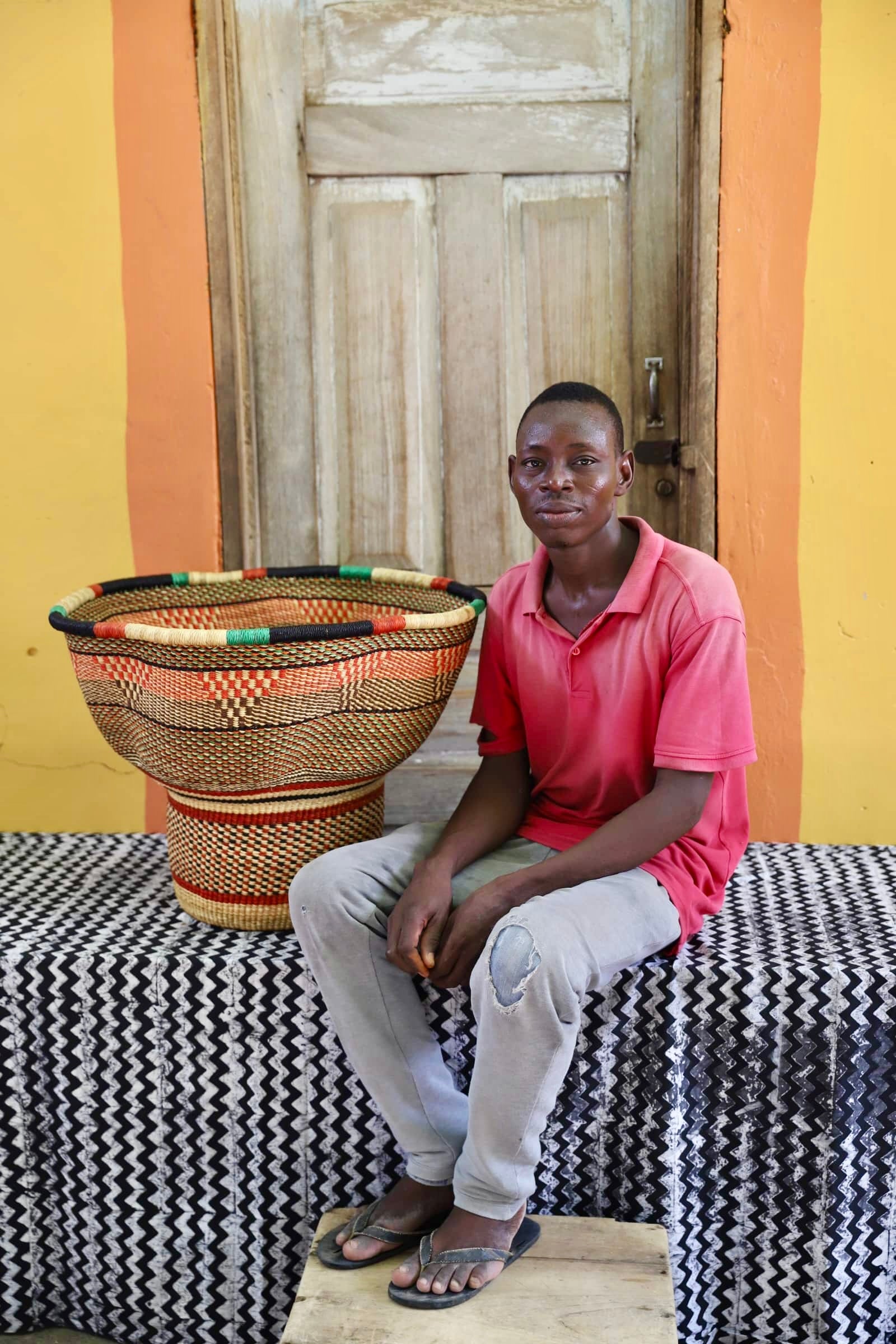 An artisan seated next to a handcrafted decorative basket with stylish straw branching out on a black and white cloth against a wooden door.
