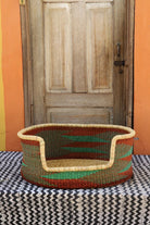 Woven pet bed with geometric pattern on a textured surface in front of a wooden door.