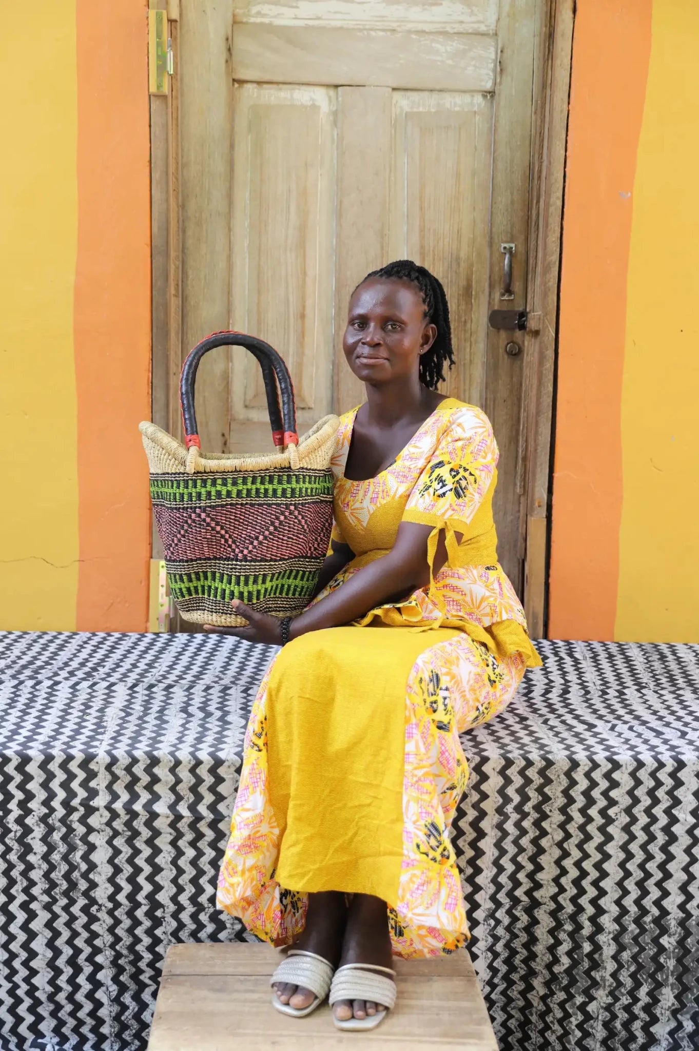 Woman in yellow dress holding a small Solara Carryall with green and dark banded stripe, leather handles